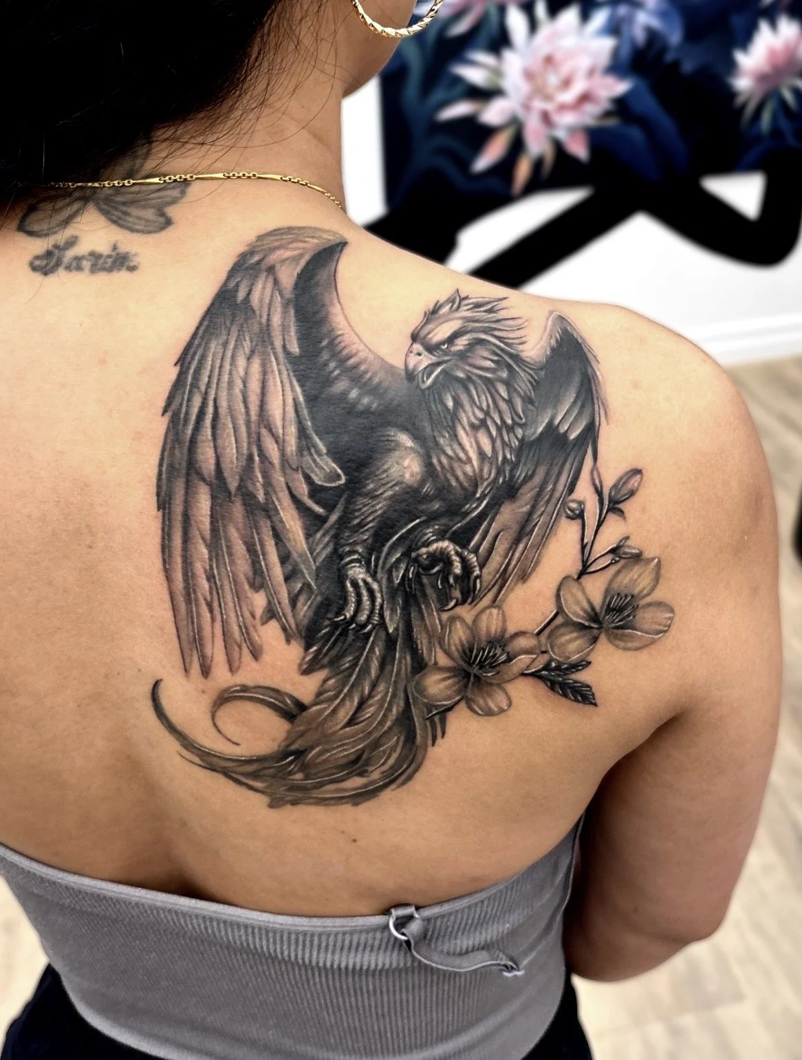 Black and grey realism tattoo o a fierce phoenix with spread wings perched on a branch with flowers on a person's shoulder.