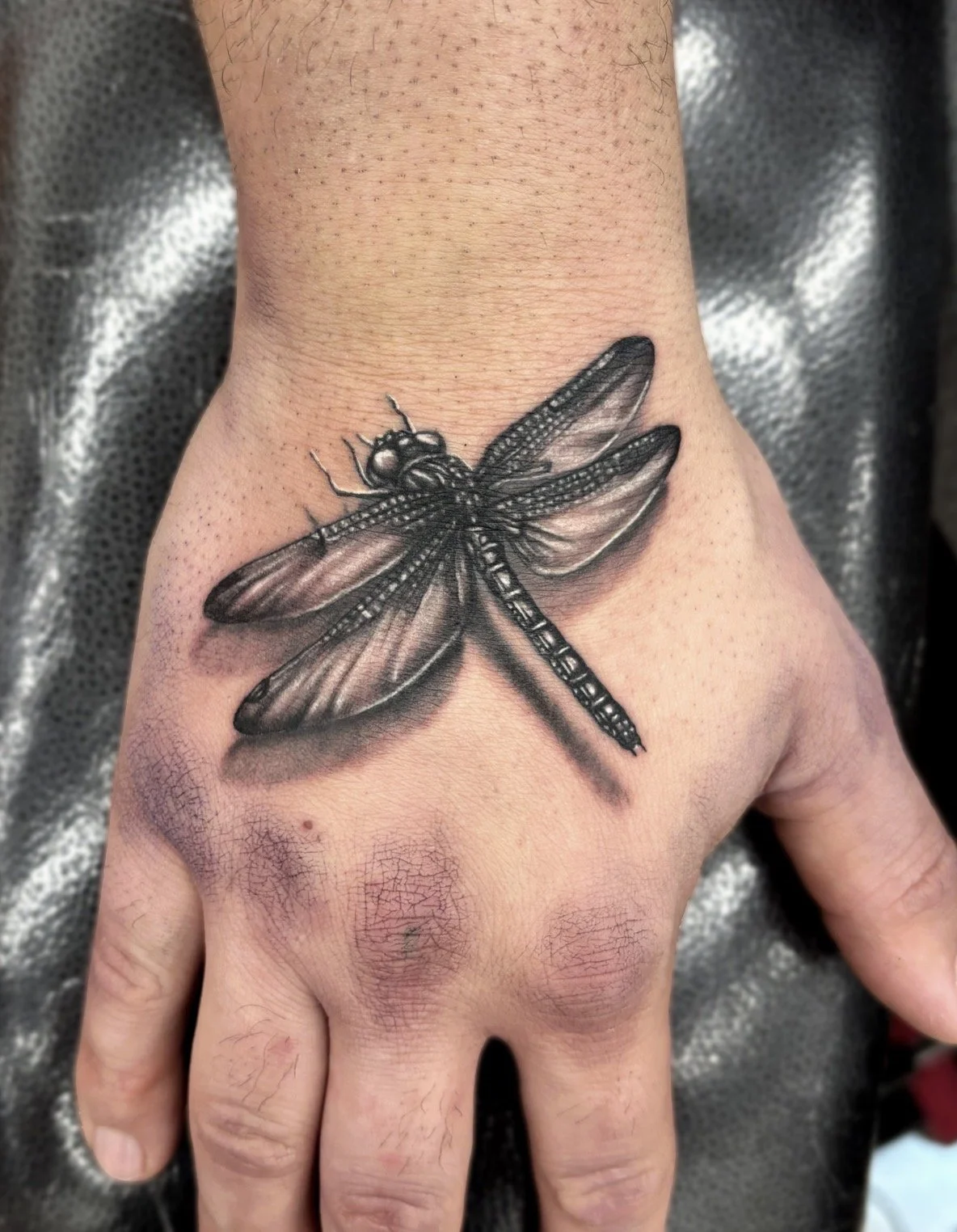 Black and grey realism tattoo of a dragonfly on the top of a person's hand.