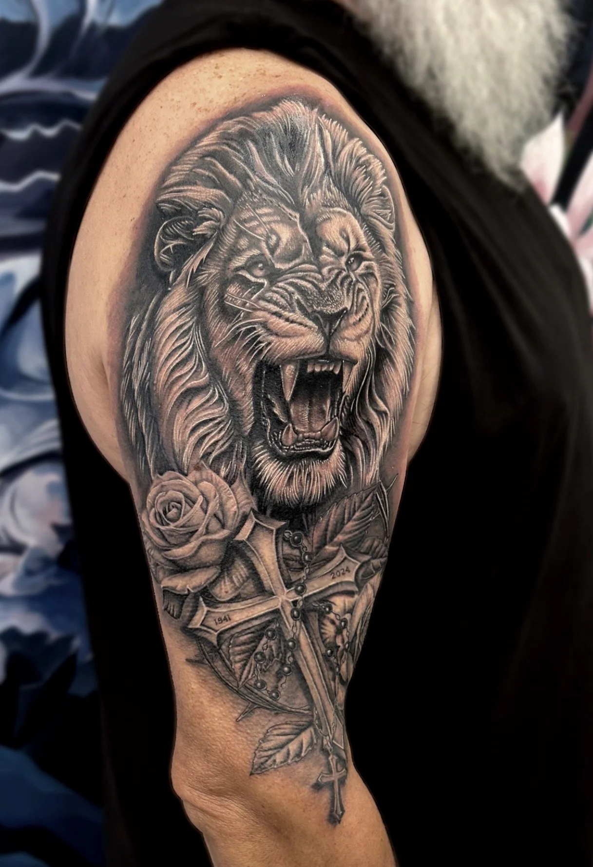 A detailed black and grey realism tattoo of a lion's head with an open mouth, a rose, and a crucifix with dates on the cross on a person's arm.