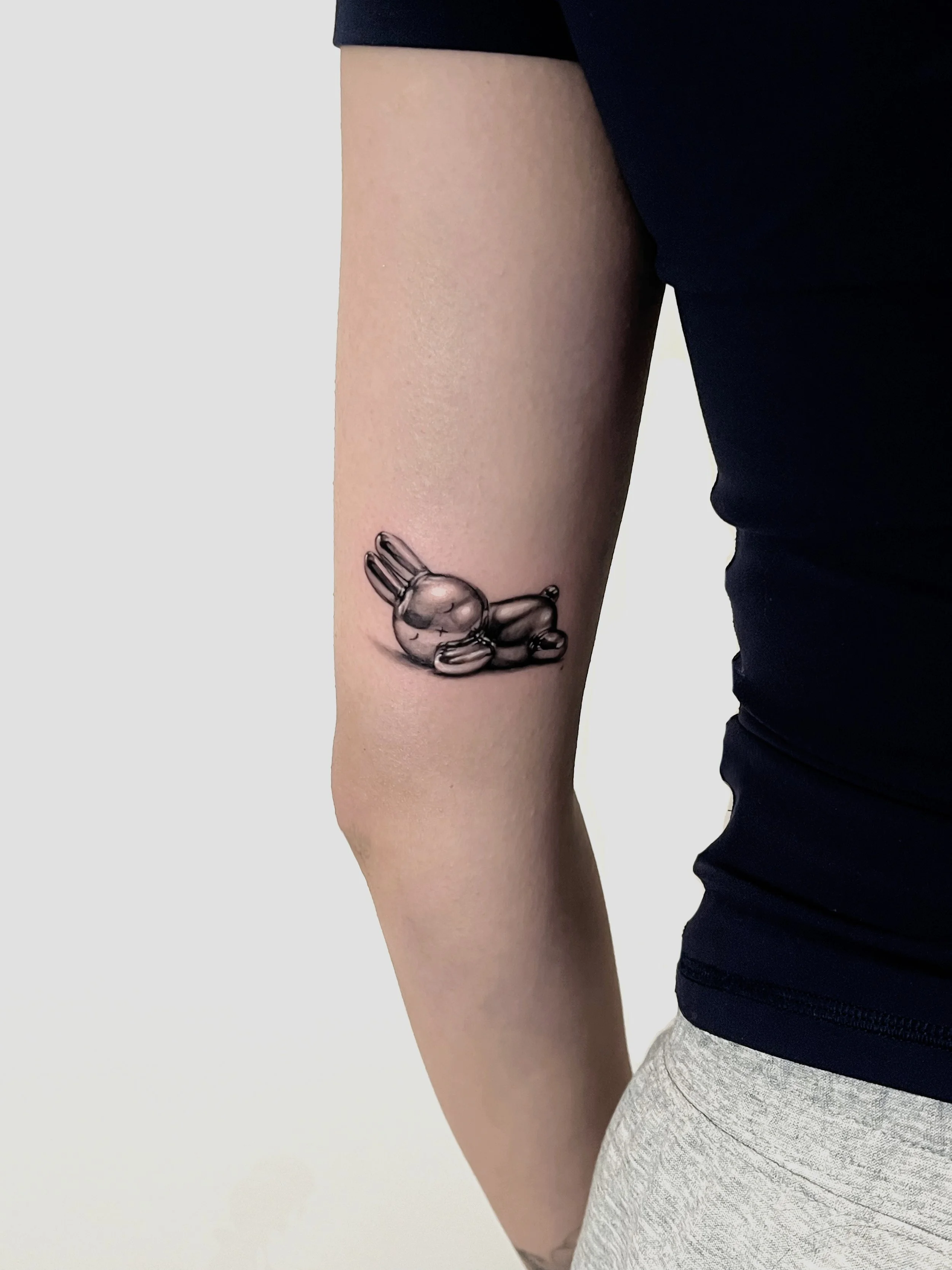 Tattoo of a metallic chrome gray bunny miffy Sanrio character lying down on the person's upper arm.