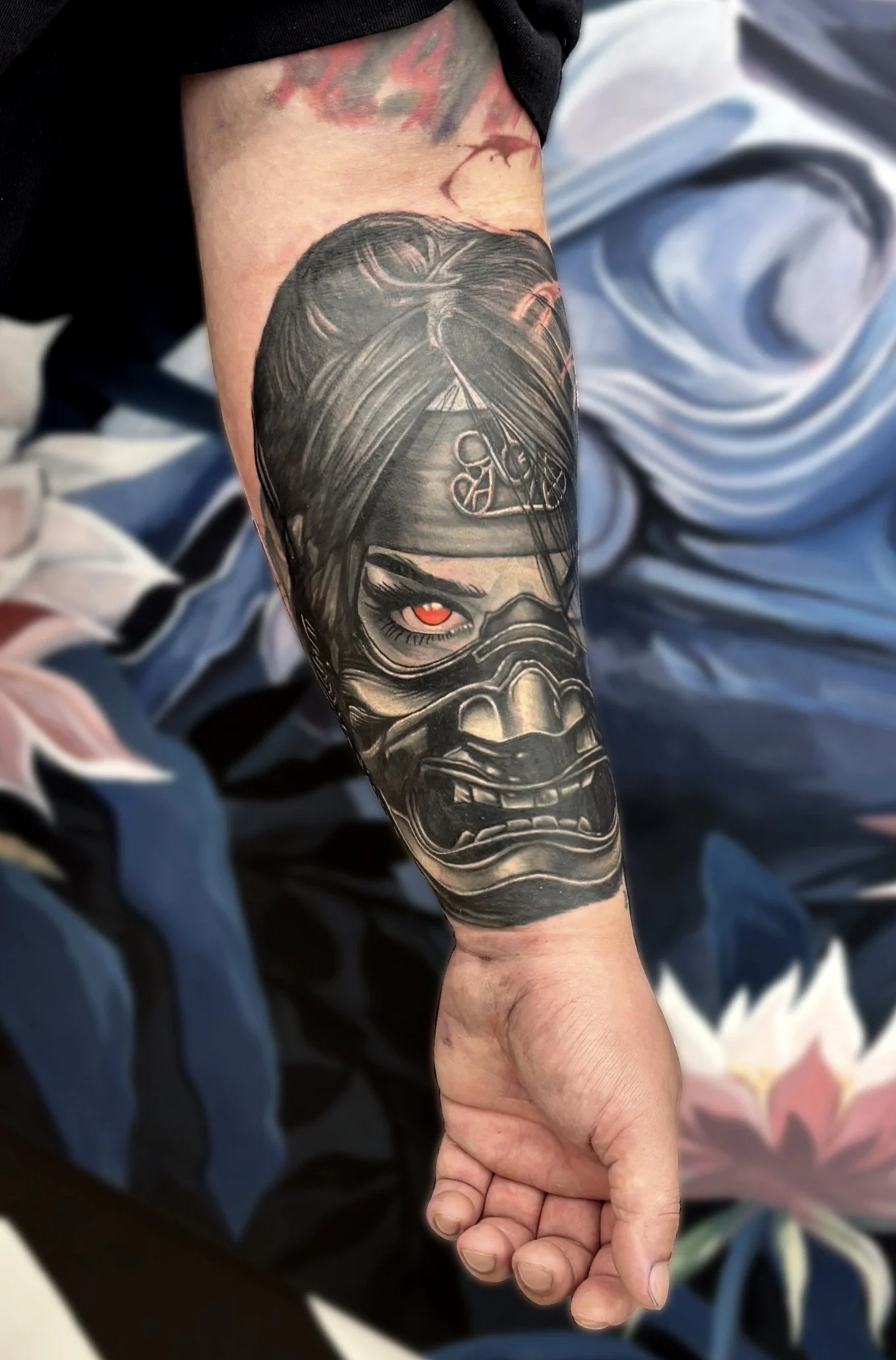 Black and grey realism tattoo of a fierce masked face/samurai with one glowing red eye on a person's forearm. This is a cover-up tattoo.