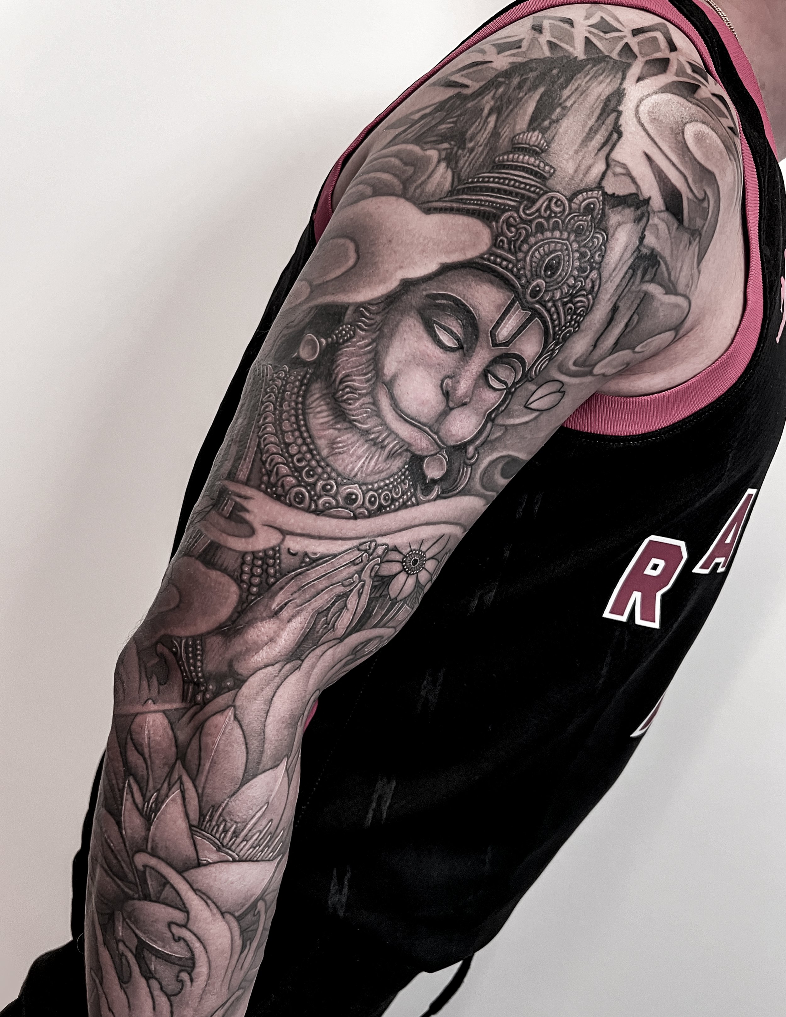 Black and grey tattoo of the Hindu God Hanuman with jewelry and a headdress, surrounded by mountains, clouds, and a lotus flower on the arm of a person.