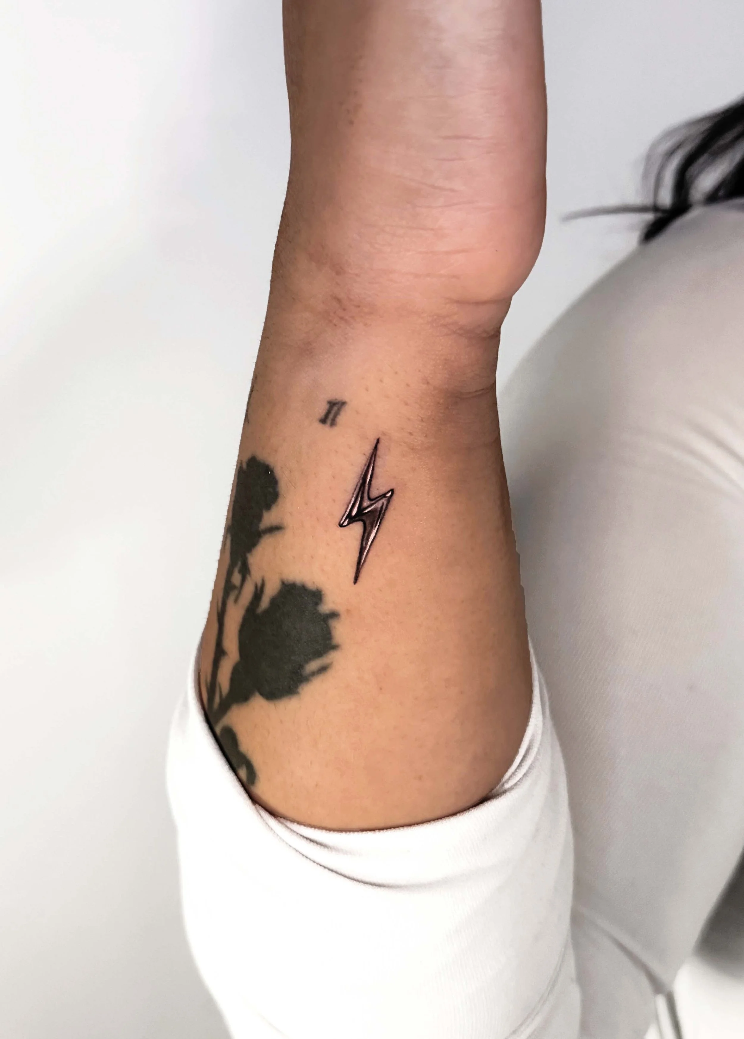 Close-up of a person's forearm with a black lightning bolt tattoo, a small Roman numeral II tattoo, and a floral tattoo, wearing a white sleeve.