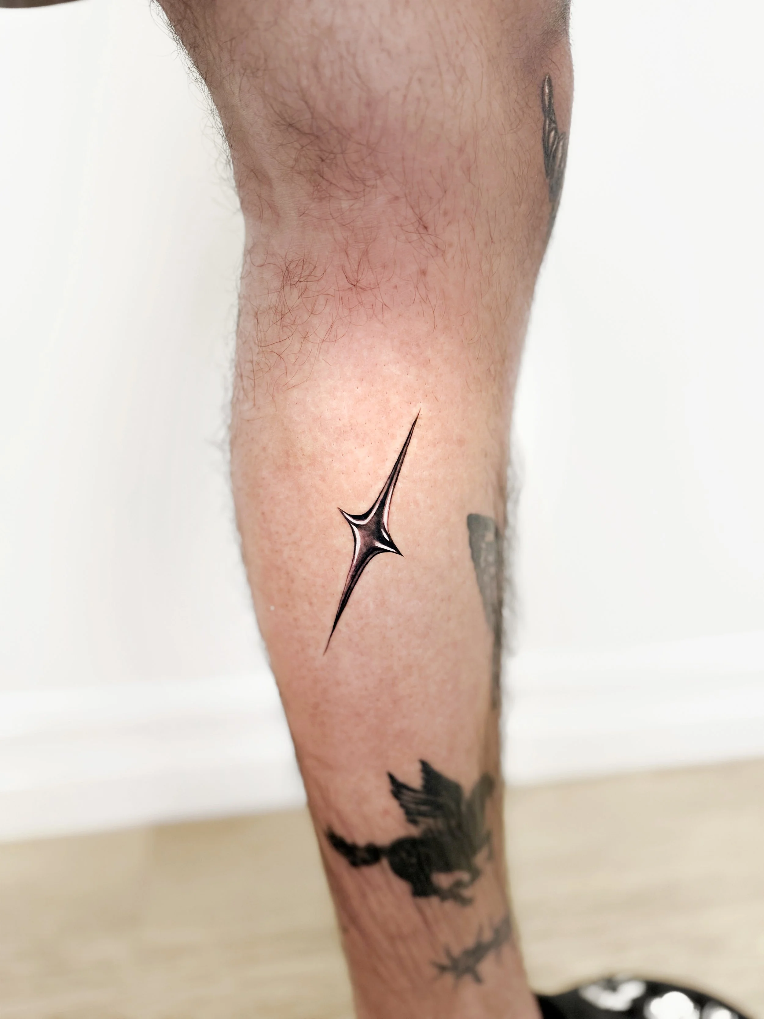Chrome tattoo of a shiny, black, stylized star on a person's calf, with additional tattoos of a bird and other designs visible on the leg.