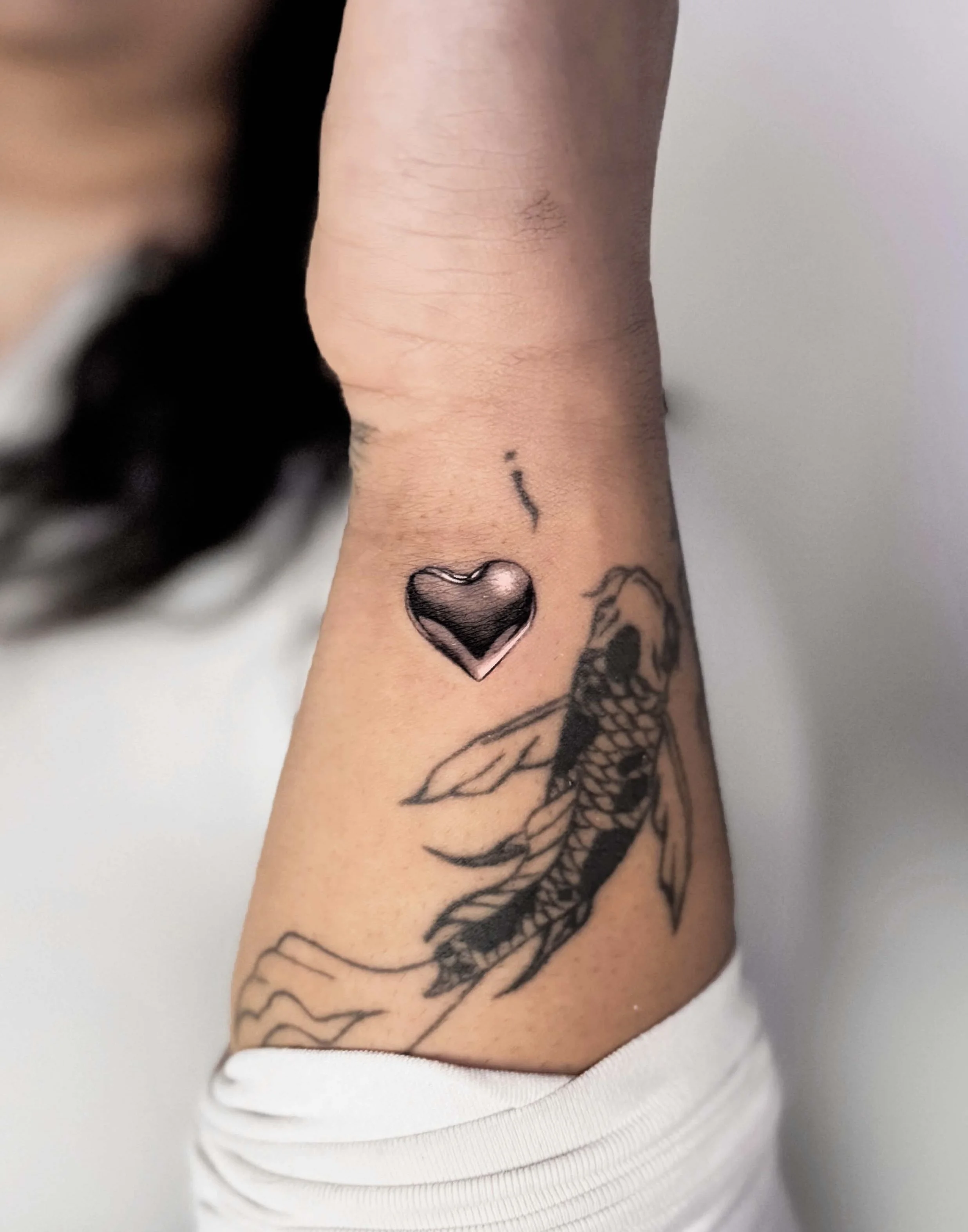 Tattoo of a fish and a chrome effect metal silver heart on a person's arm.
