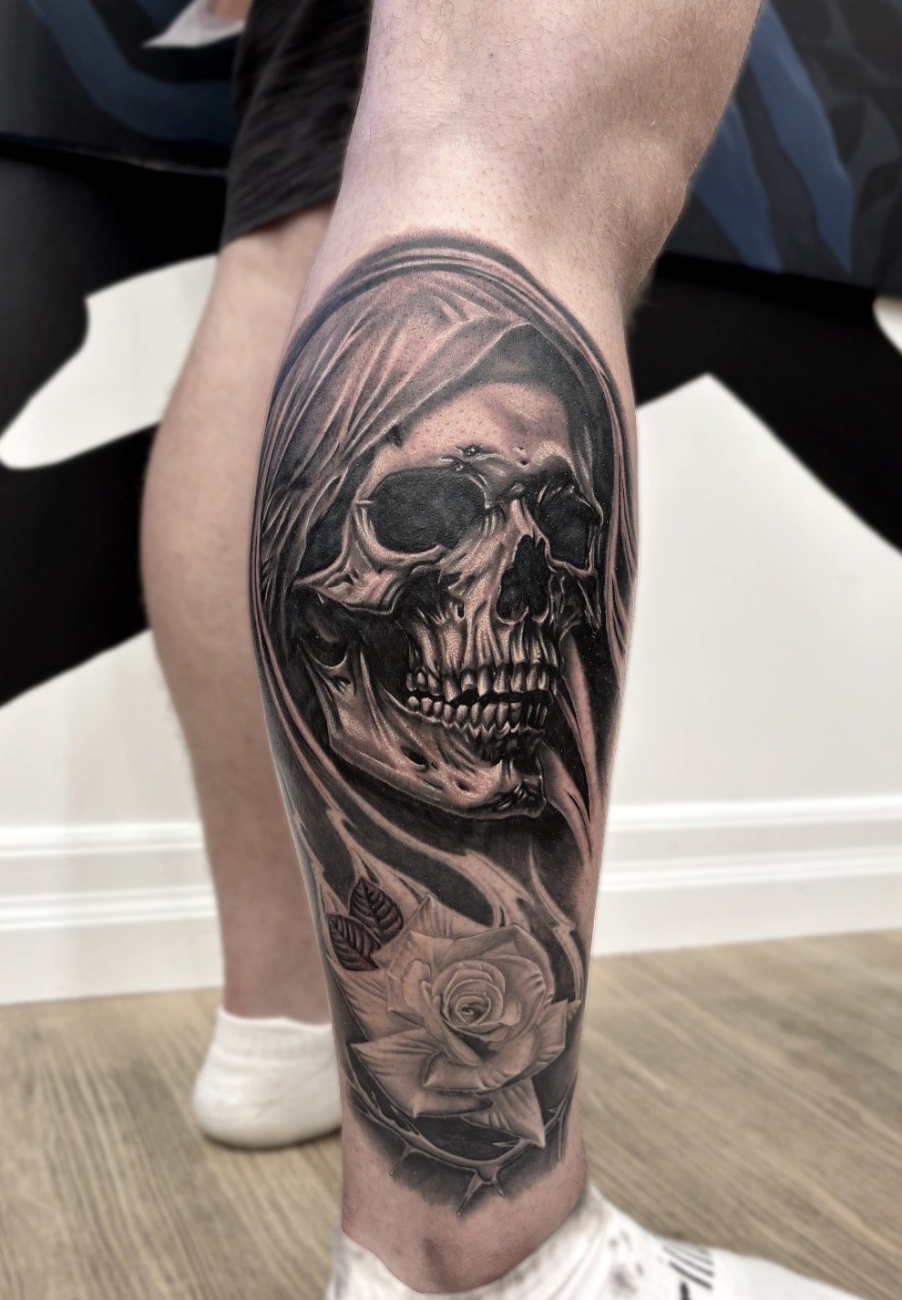A detailed black and grey tattoo of a skull with a hooded figure and a rose on a person's calf.