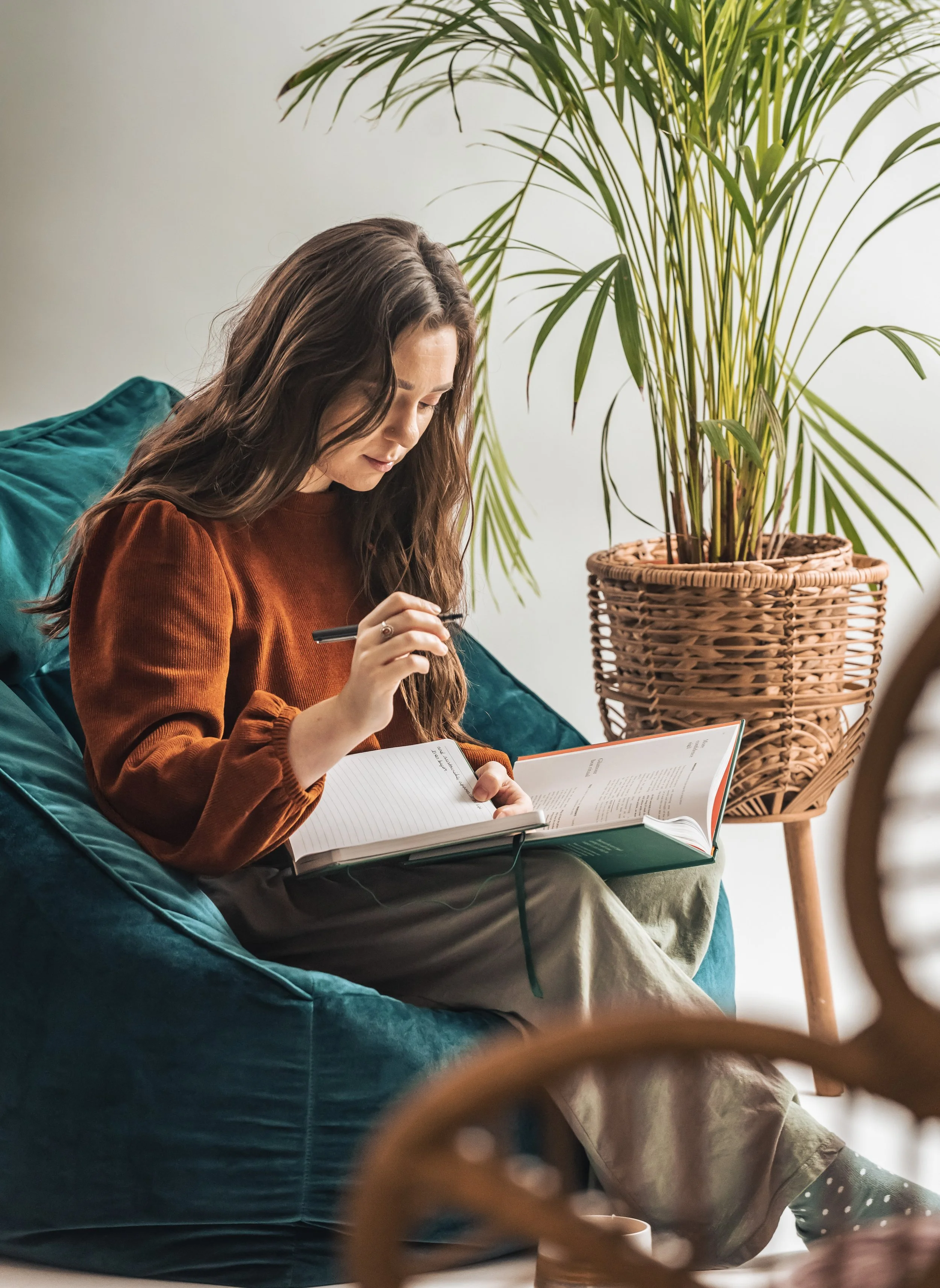 A woman with long wavy brown hair sitting on a teal velvet cushion on a sofa, reading a book and holding a pen in her right hand, with a large potted plant with long green leaves in the background.