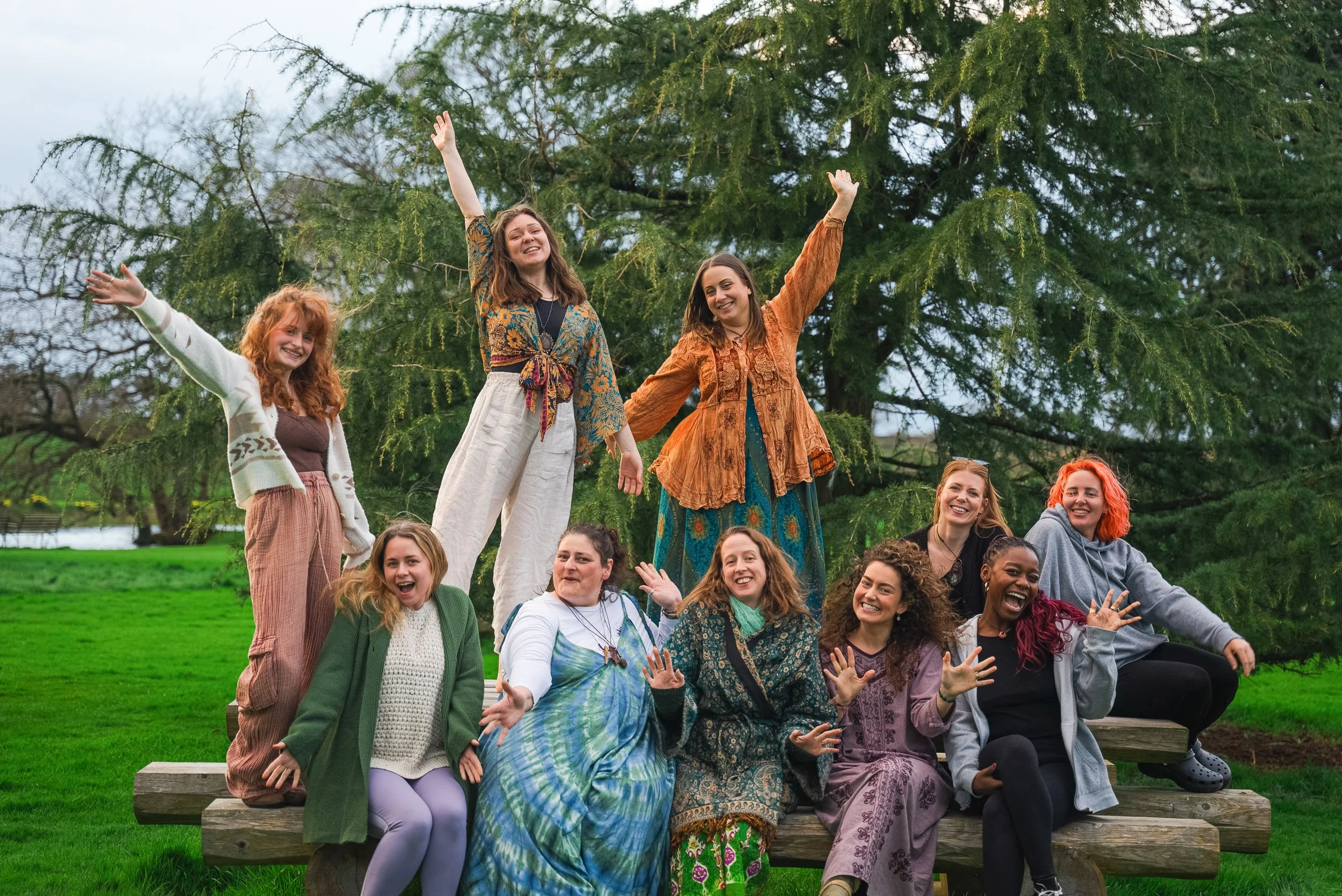 A group of eleven women smiling and cheering outdoors in front of a large tree, some sitting on a wooden bench and others standing behind it, celebrating together on a grassy lawn.