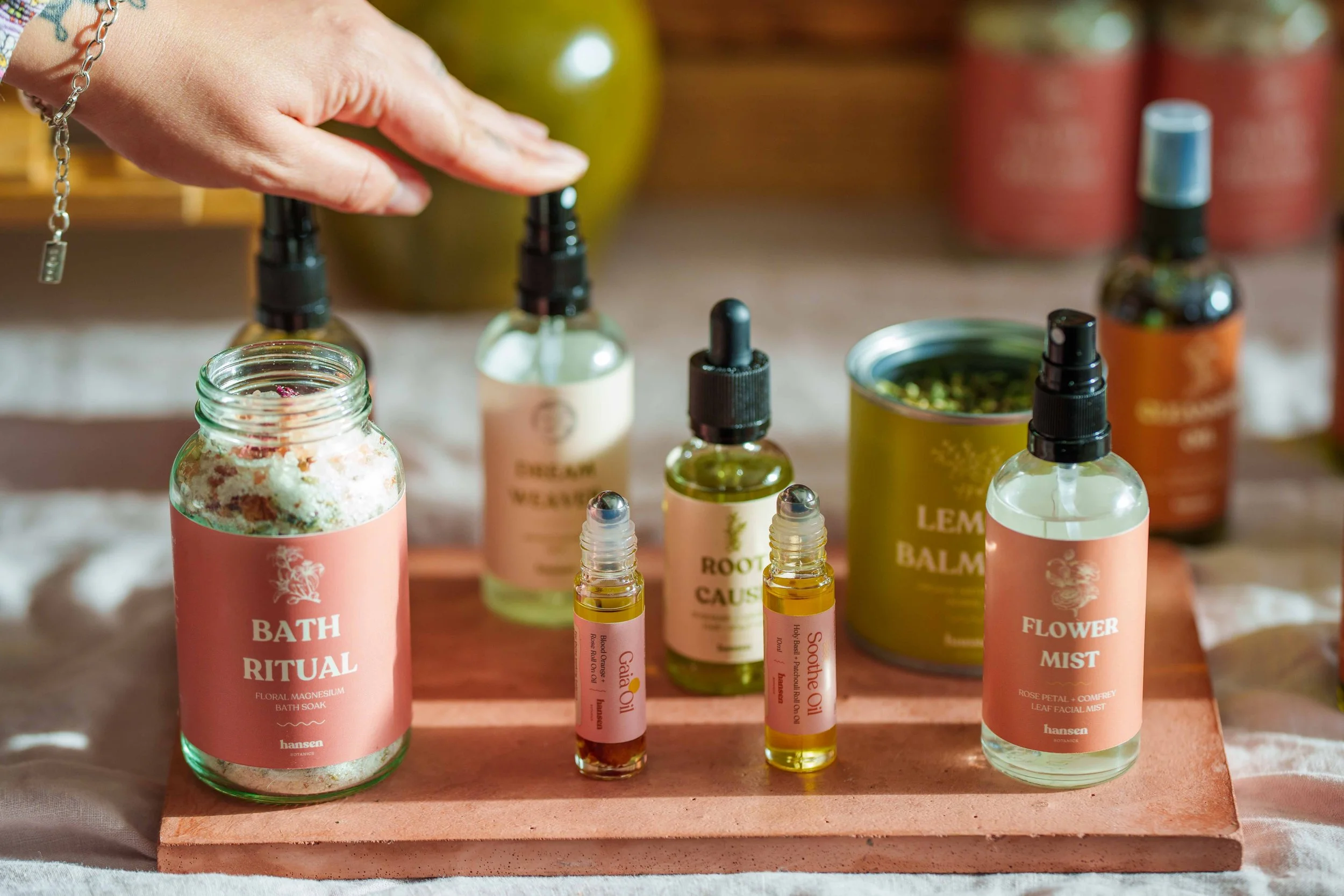 Assorted essential oils and bath products displayed on a pink wooden board, including bath soak, room spray, bath balm, and flower mist, with some products in spray bottles and others in jars and small bottles.
