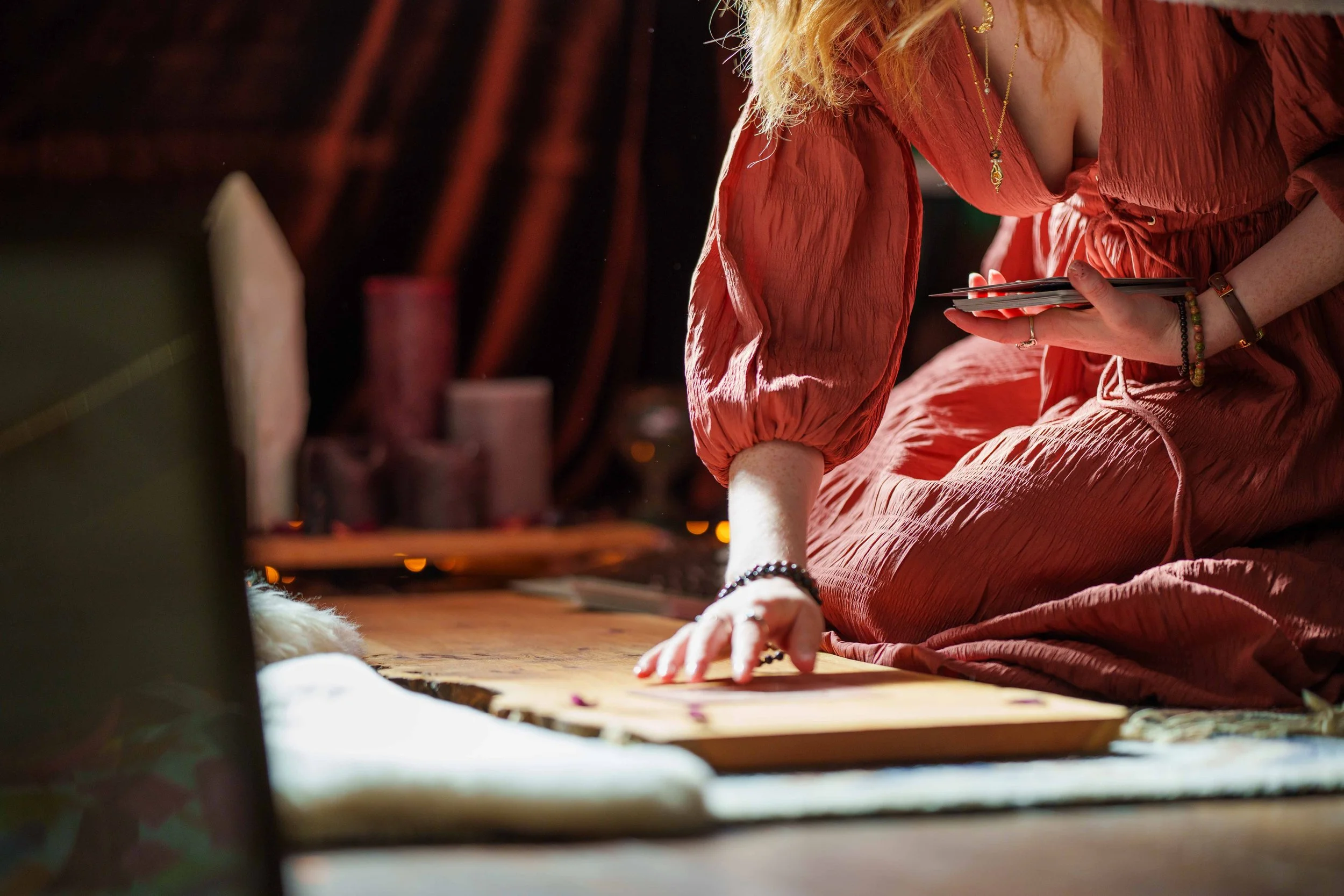 A woman in a reddish-orange dress is sitting on the ground, leaning forward with one hand on a wooden surface. She is holding a few cards in her other hand and wearing jewelry, including a necklace, bracelets, and rings. The background is dark and ou