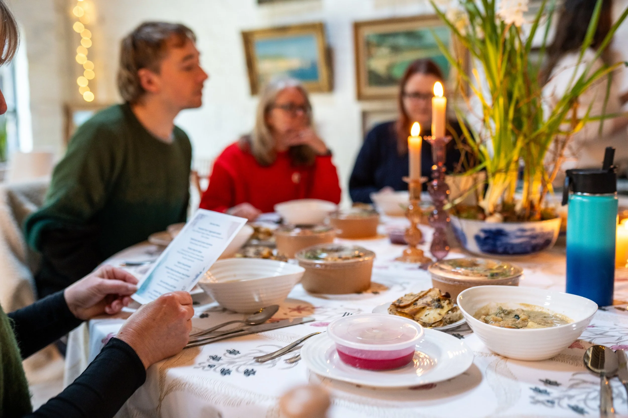 Retreat guests gathered around a dinner table with food, candles, and decorations, with several paintings on the wall in the background.