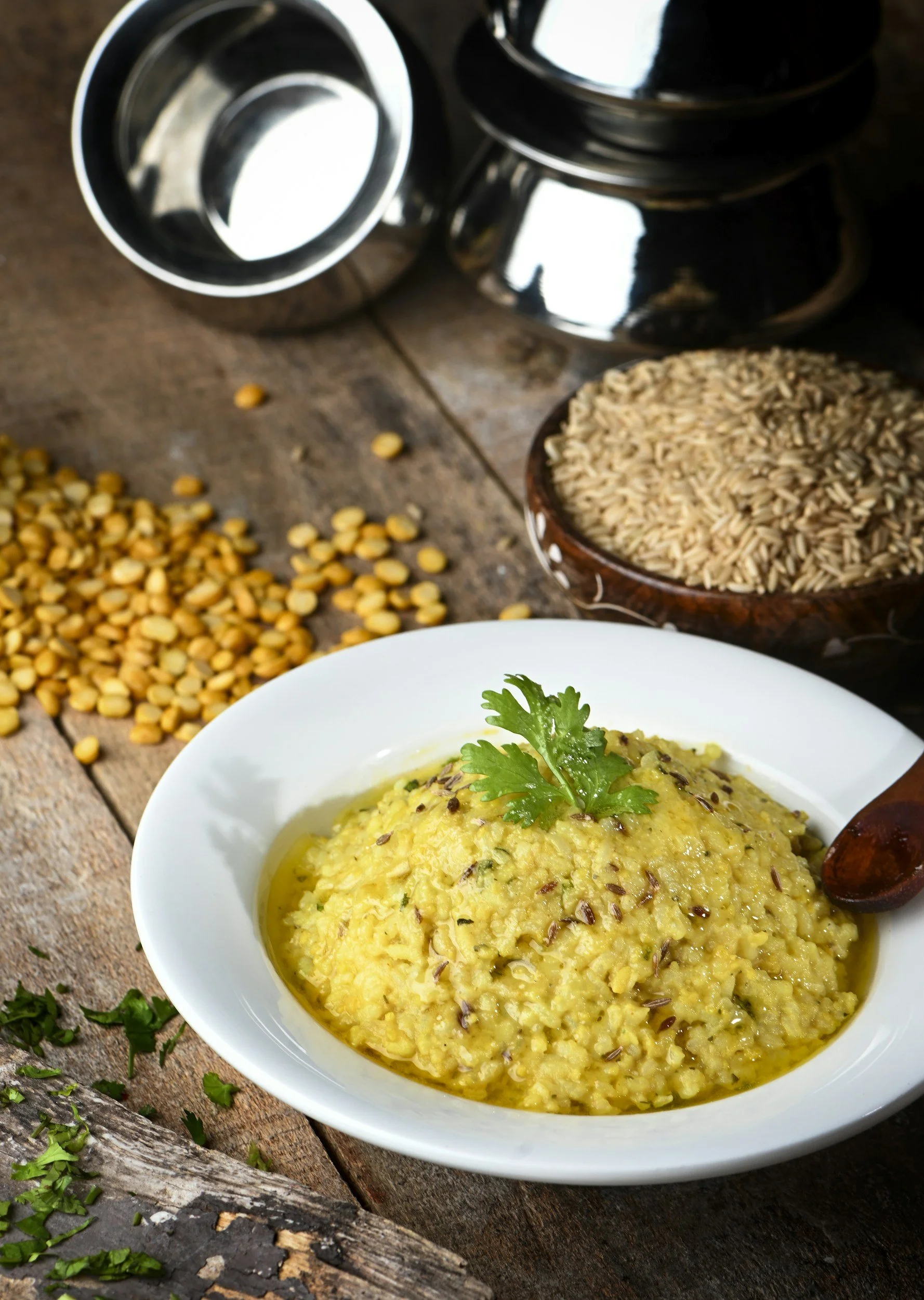 A bowl of yellow lentil dal garnished with cilantro, surrounded by dried lentils and grains on a rustic wooden surface.