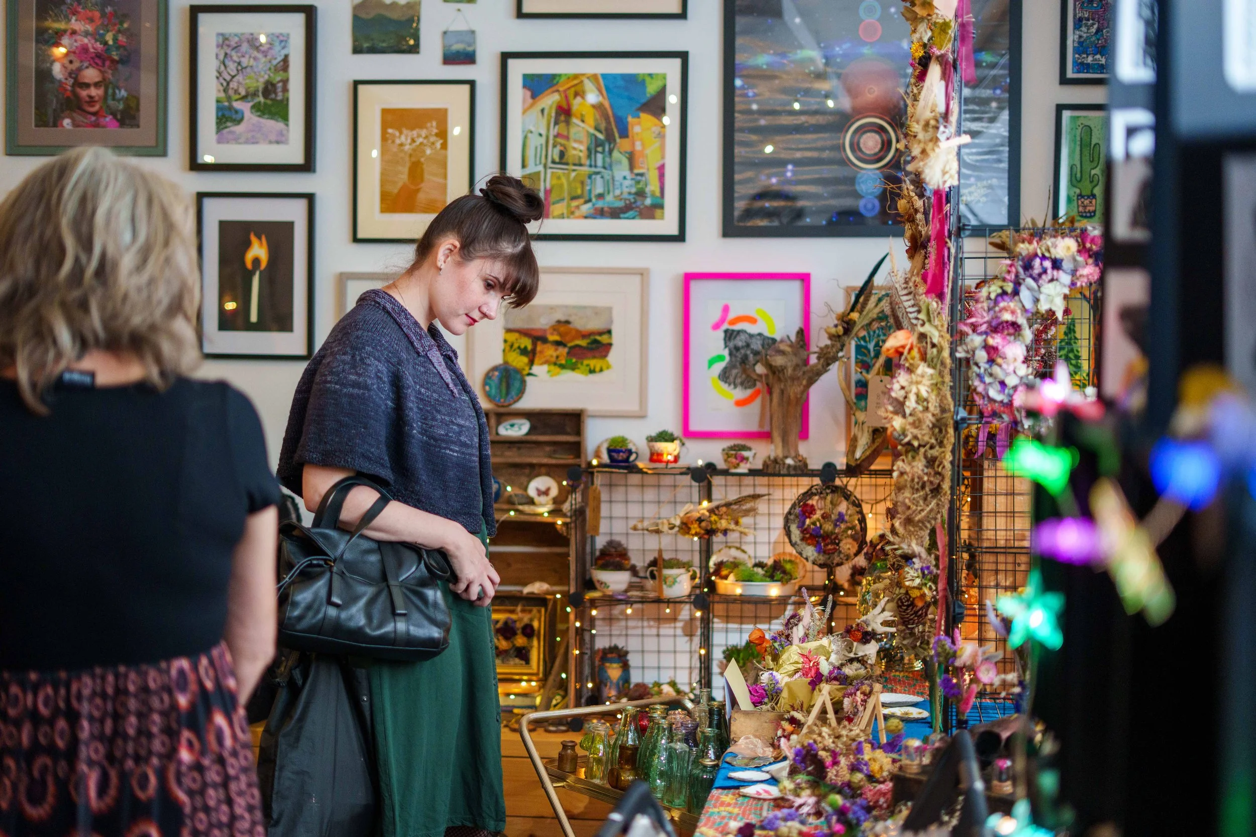 Two women shopping at an art and craft fair. One woman with blonde hair and a black shirt is looking at a table with small bottles and decorative items. The other woman, with brown hair in a bun and wearing a dark jacket, is examining the merchandise