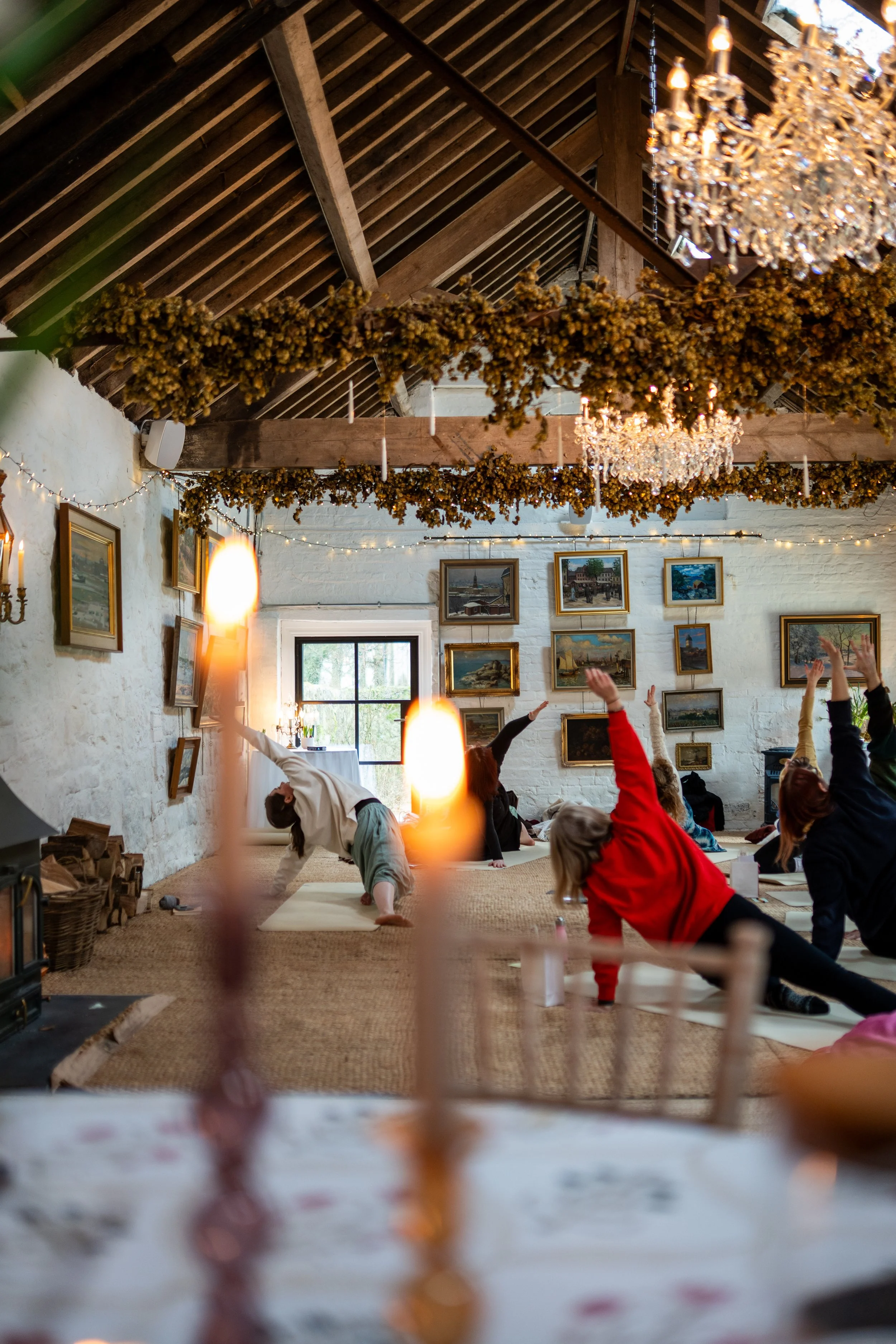 People participating in a yoga class in a cozy, rustic studio with white brick walls, framed paintings, chandeliers, and hanging dried flowers.