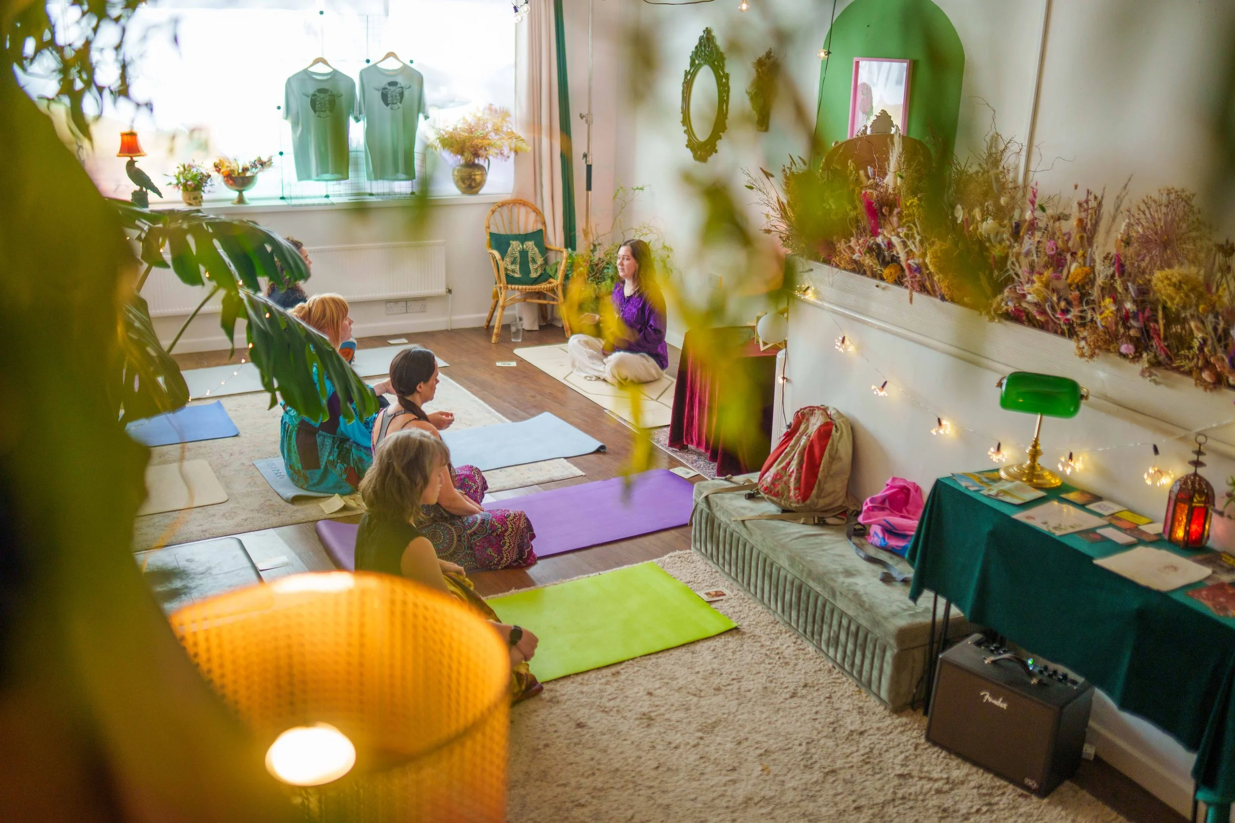 Women leading a yoga class in a cosy, decorated room with mats on the floor, flowers, and vintage decor.