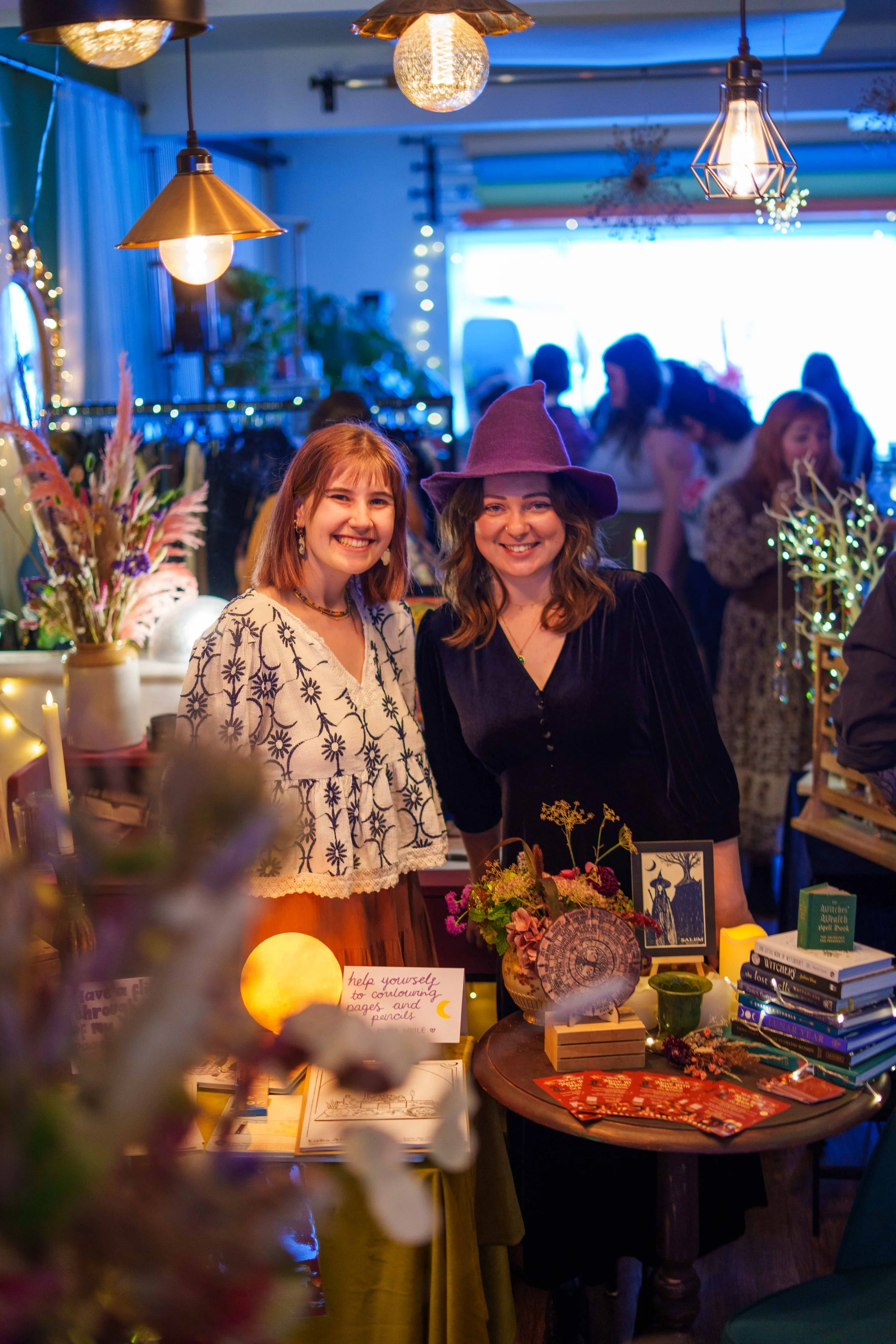 Two women smiling at a booth with books and decorations, one wearing a purple witch hat, at a social gathering inside a decorated room.