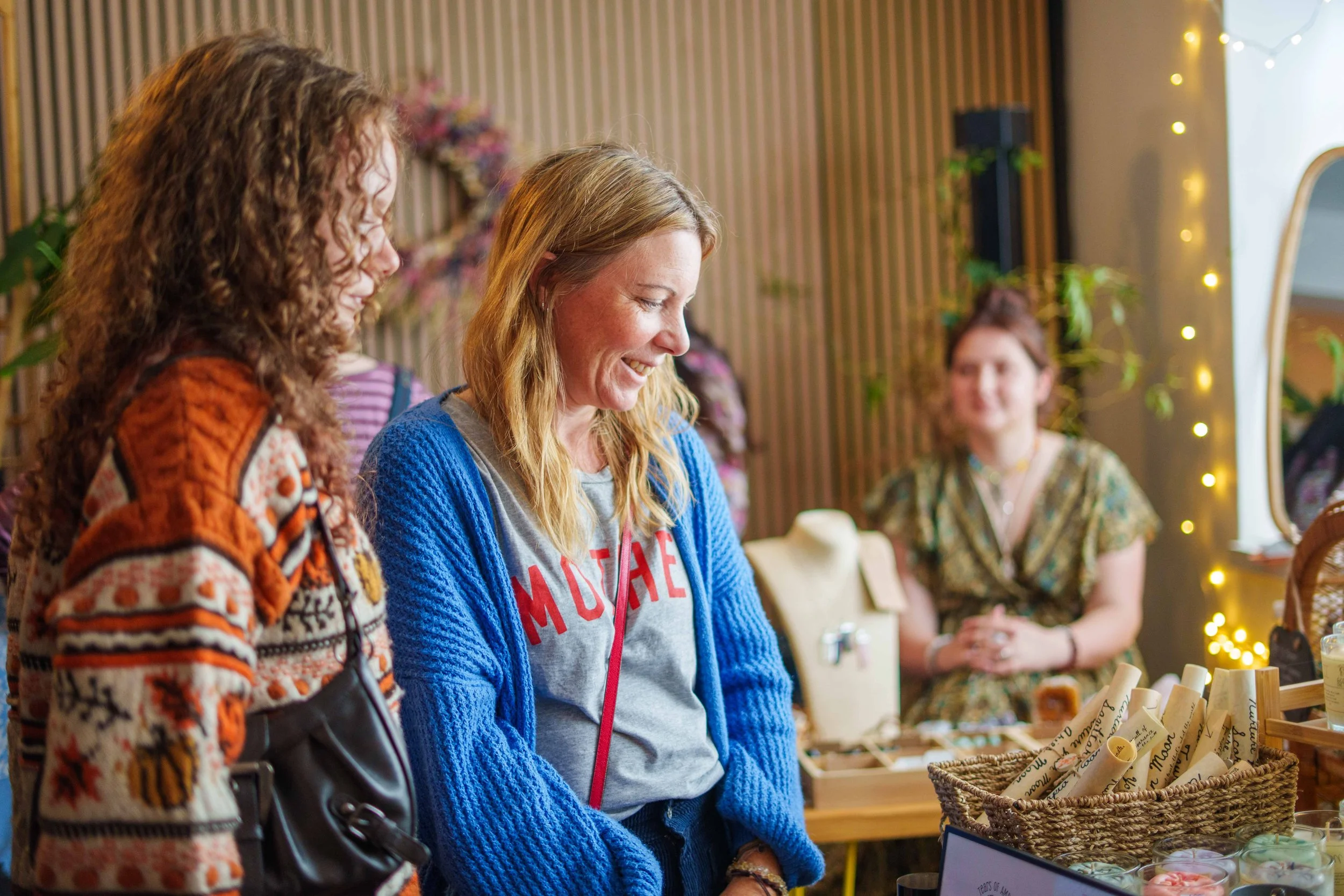 Three women smiling and browsing at a market stall inside a decorated shop.