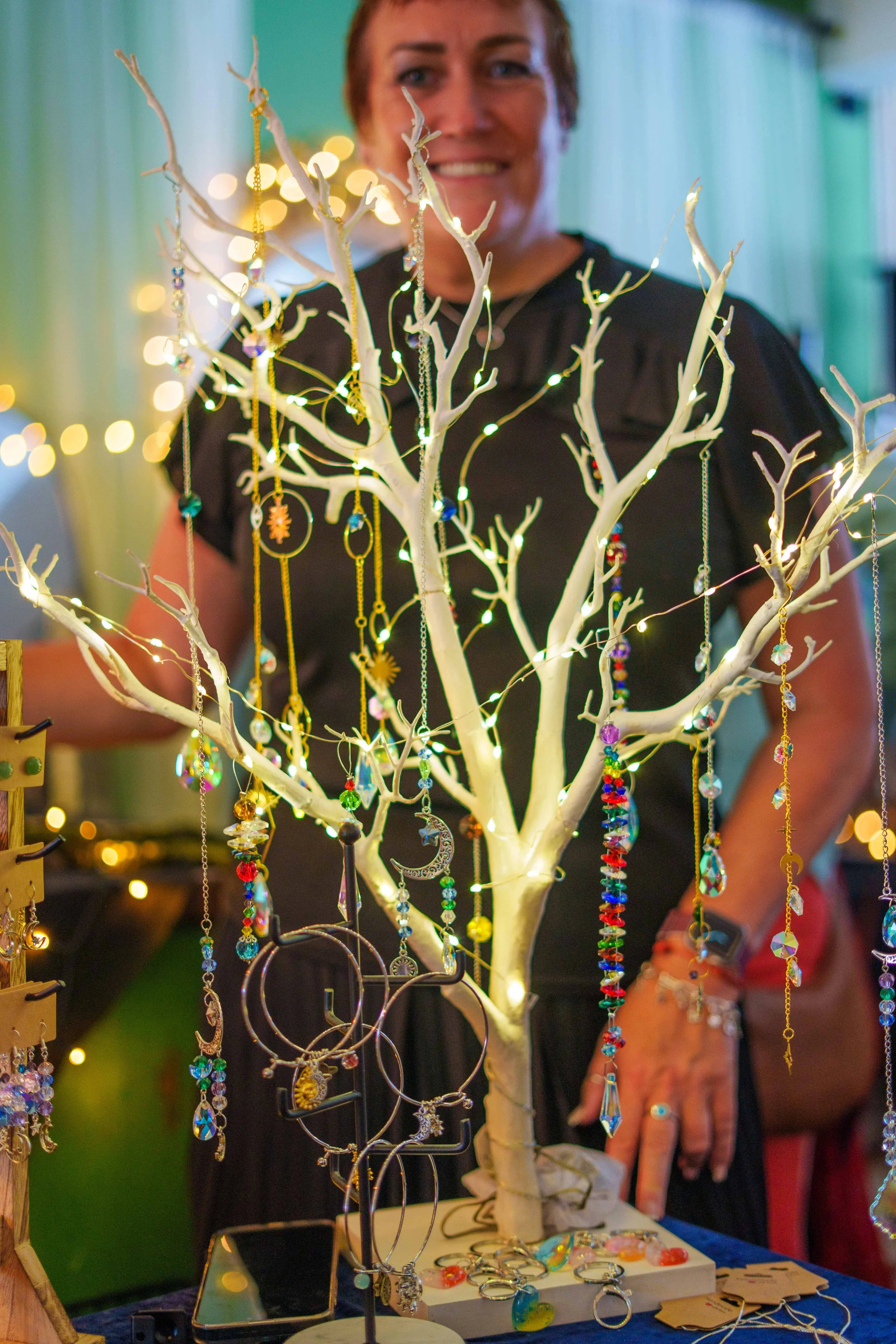 A woman smiling behind a white decorative tree displaying colorful jewelry, including necklaces, earrings, and rings, with a blurred background of string lights.