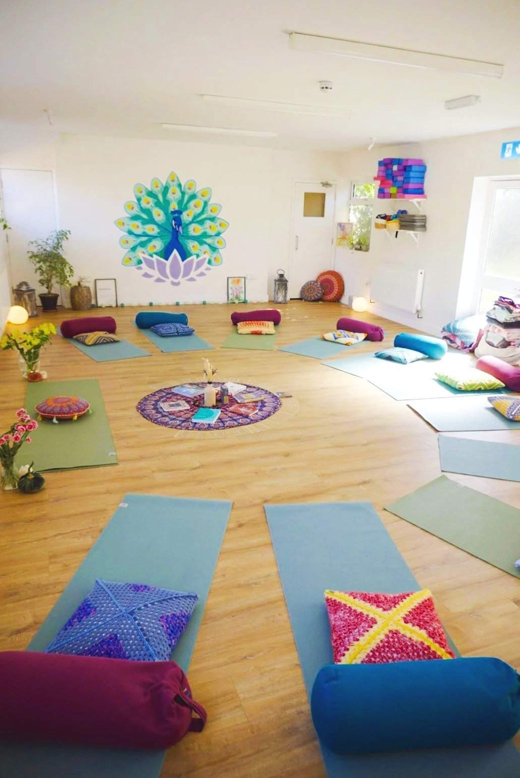 A spacious yoga room with wooden floor, colourful cushions, mats, and props arranged in a circle. A peacock mural decorates the white wall, with plants and decorative items around.