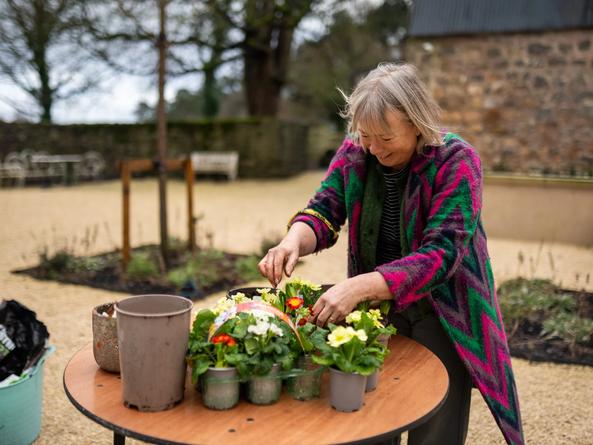 A woman in a colorful jacket planting flowers on a round wooden table outdoors.