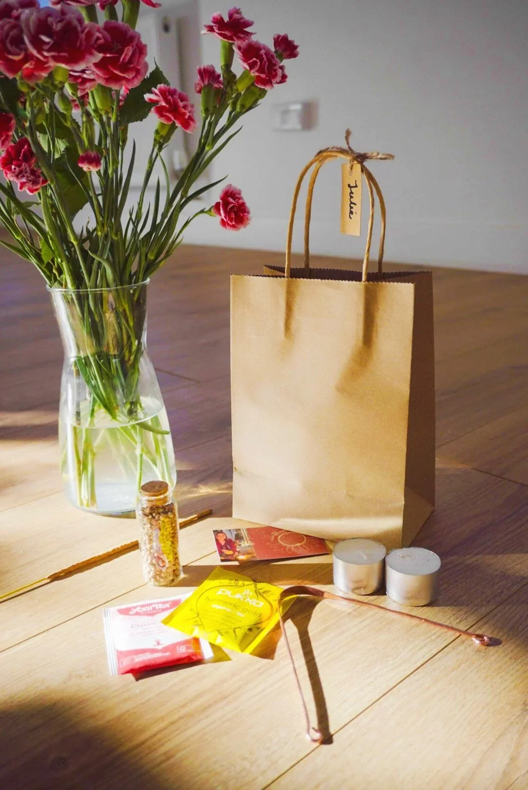 A wooden table with pink carnations in a glass vase, a small bottle of seeds, a yellow tea bag, a red tea bag, two tealight candles, and a copper metal tongue scraper, with a blank brown paper bag and a white wall in the background.