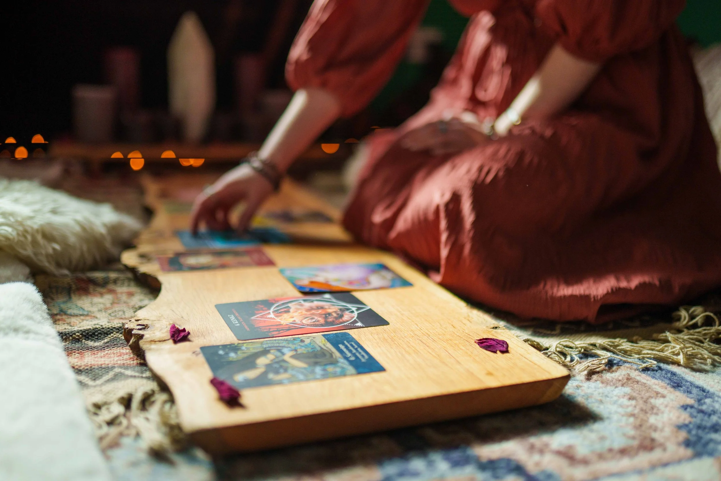 Person in a red dress sitting on the floor, reaching out to tarot cards laid on a wooden board with a patterned rug underneath, outdoors at night with city lights in the background.