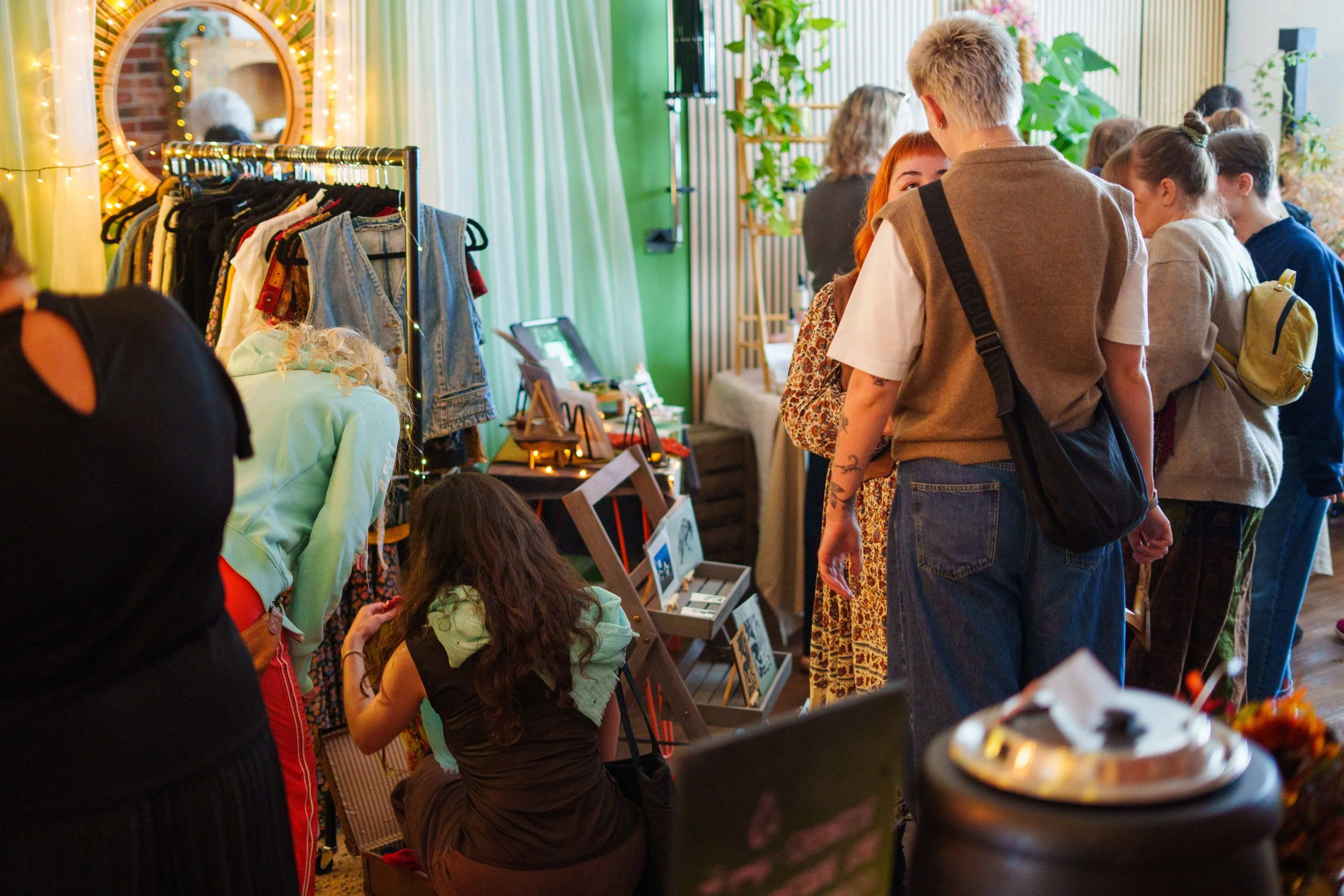 People browsing a pop-up shop or market with clothing, jewelry, and decorative items displayed on tables and racks, in a cozy, well-lit indoor space with green and white curtains, plants, and a mirror with fairy lights.
