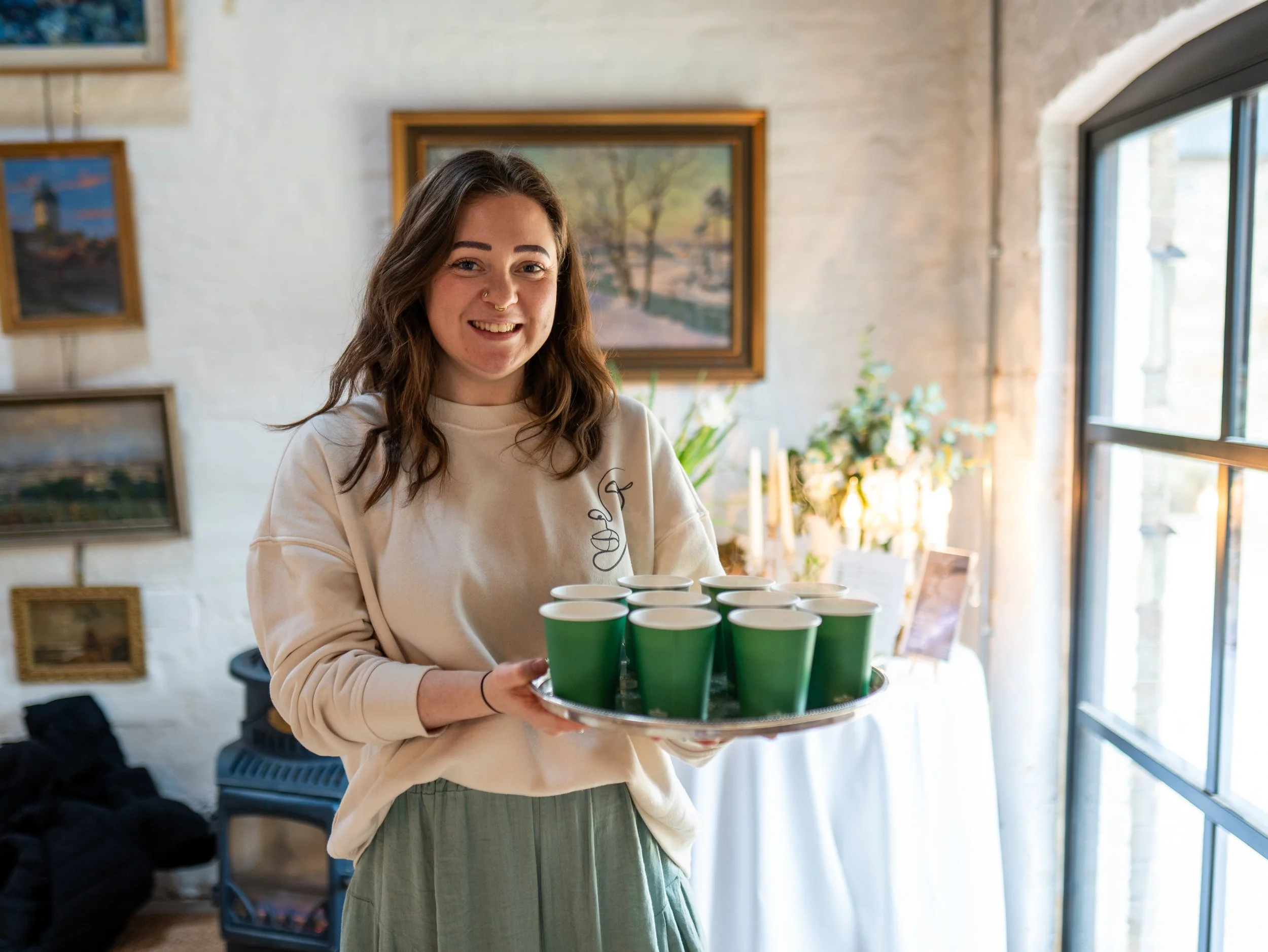 A young woman with brown hair smiling and holding a tray of green paper cups in a well-lit indoor space with framed paintings and large windows.