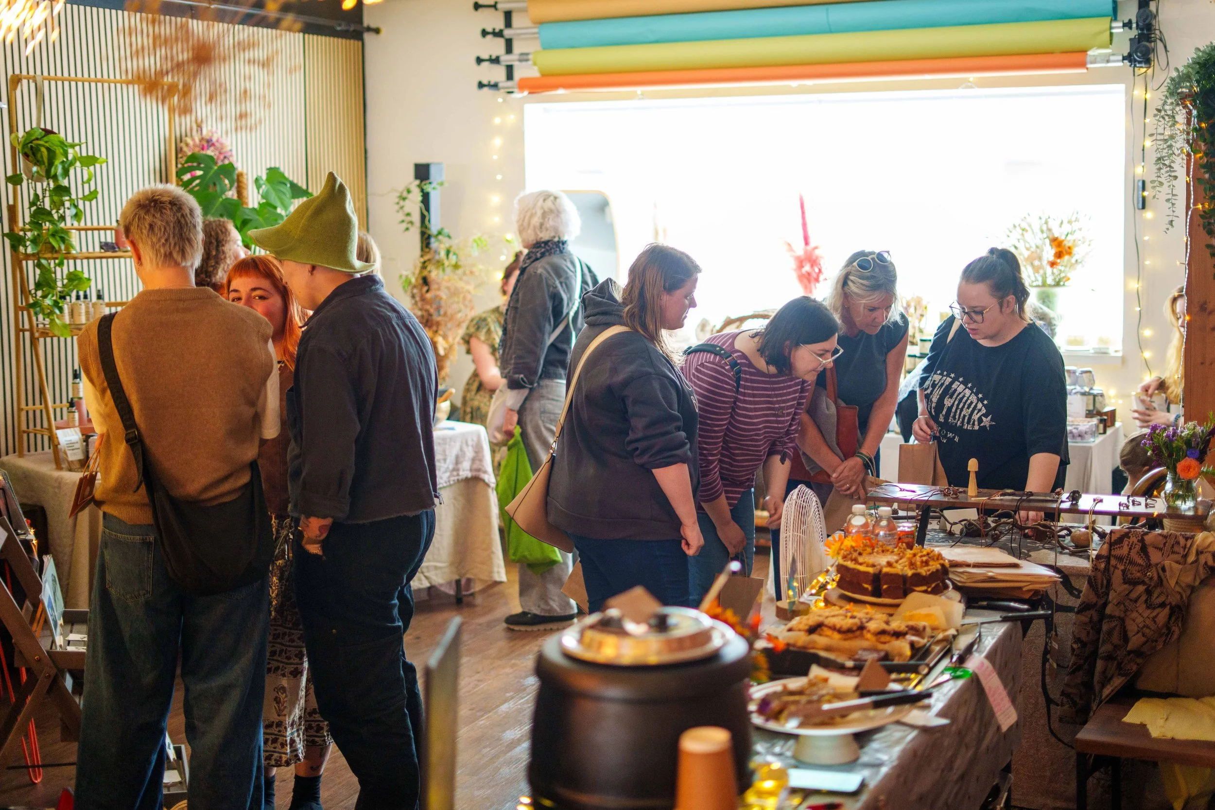People shopping at a craft fair or market with tables displaying baked goods and handmade items, decorated with string lights, plants, and colorful background elements.