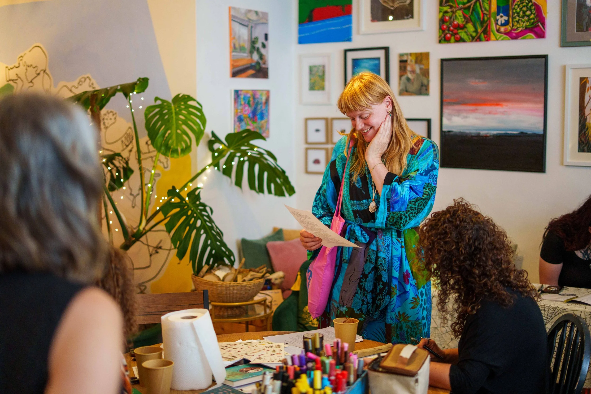 A woman with red hair, wearing a colorful blue patterned dress, is smiling and reading a piece of paper at a table. She has a pink and gray bag on her shoulder. Others are seated around the table, which has art supplies and paper on it. The backgroun