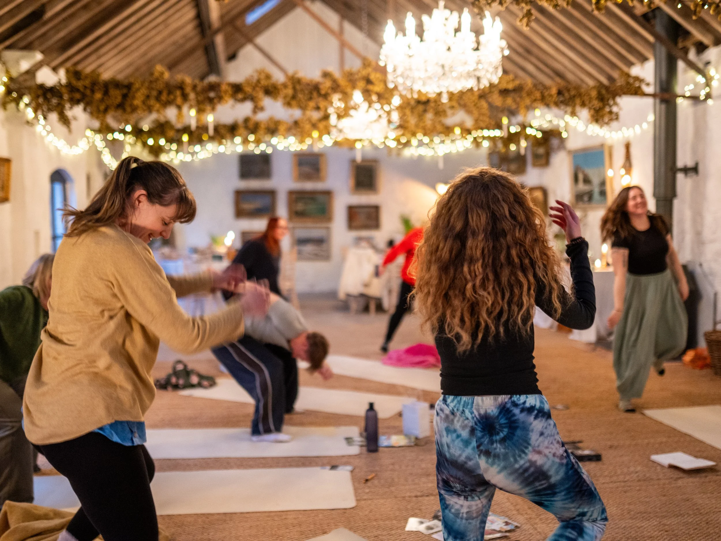 Women practicing yoga or dancing in a decorated indoor space with string lights, a chandelier, and framed pictures on the wall.