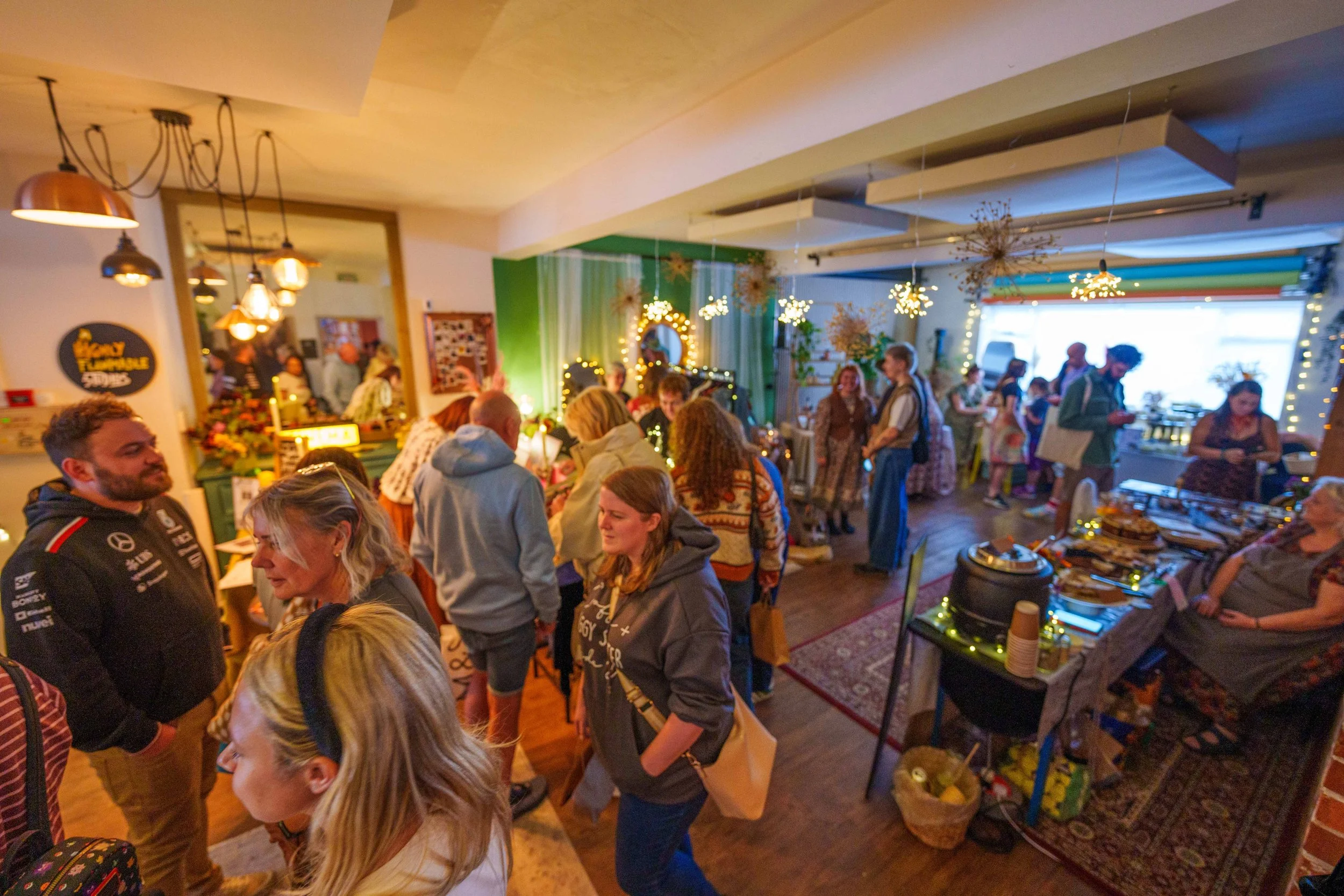 People gathered in a decorated indoor space with string lights, floral arrangements, and colorful decorations, likely at a social event or market.