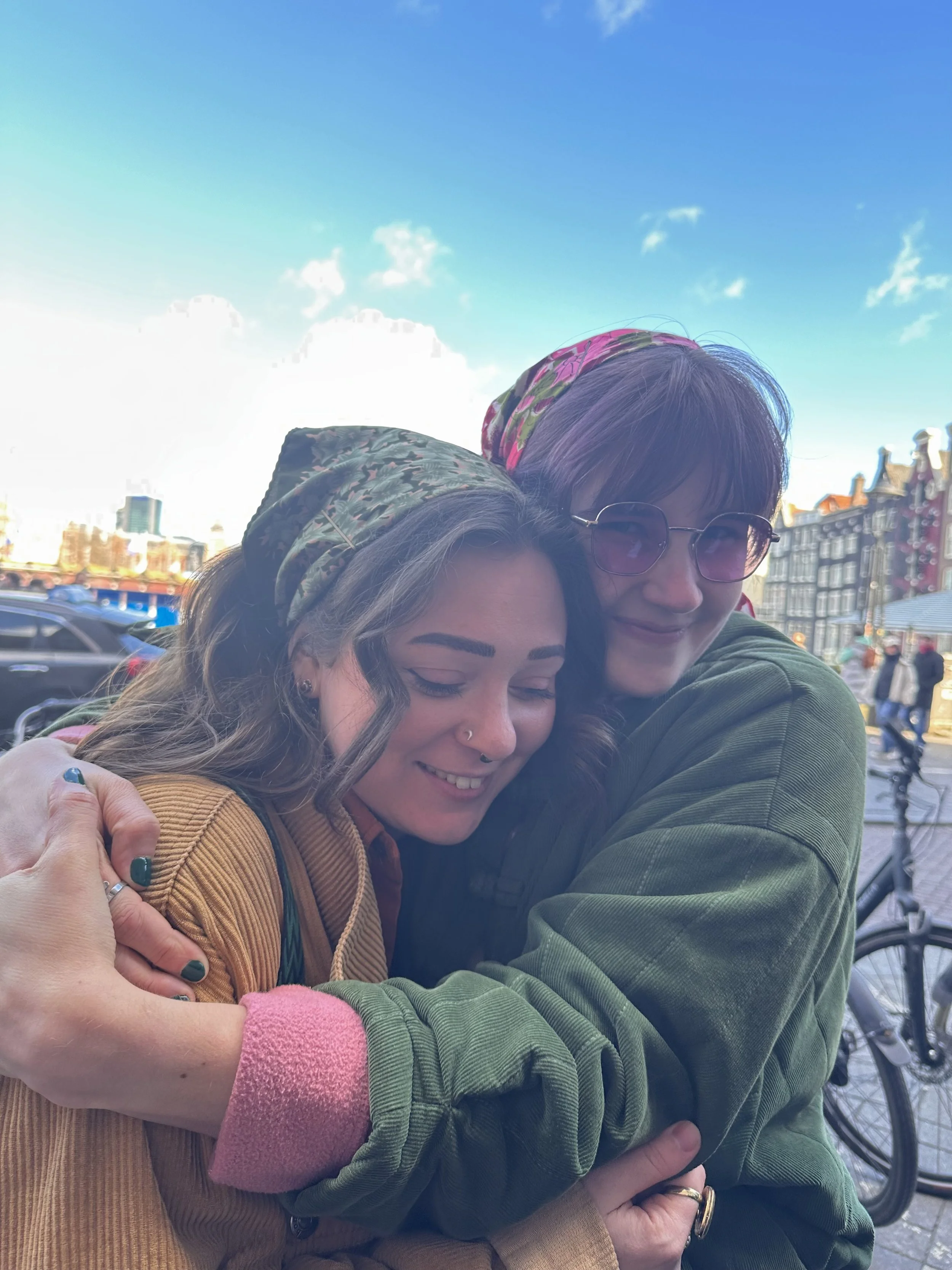 Two women hugging each other with smiles on their faces, outdoors in an urban setting with bicycles and buildings in the background.