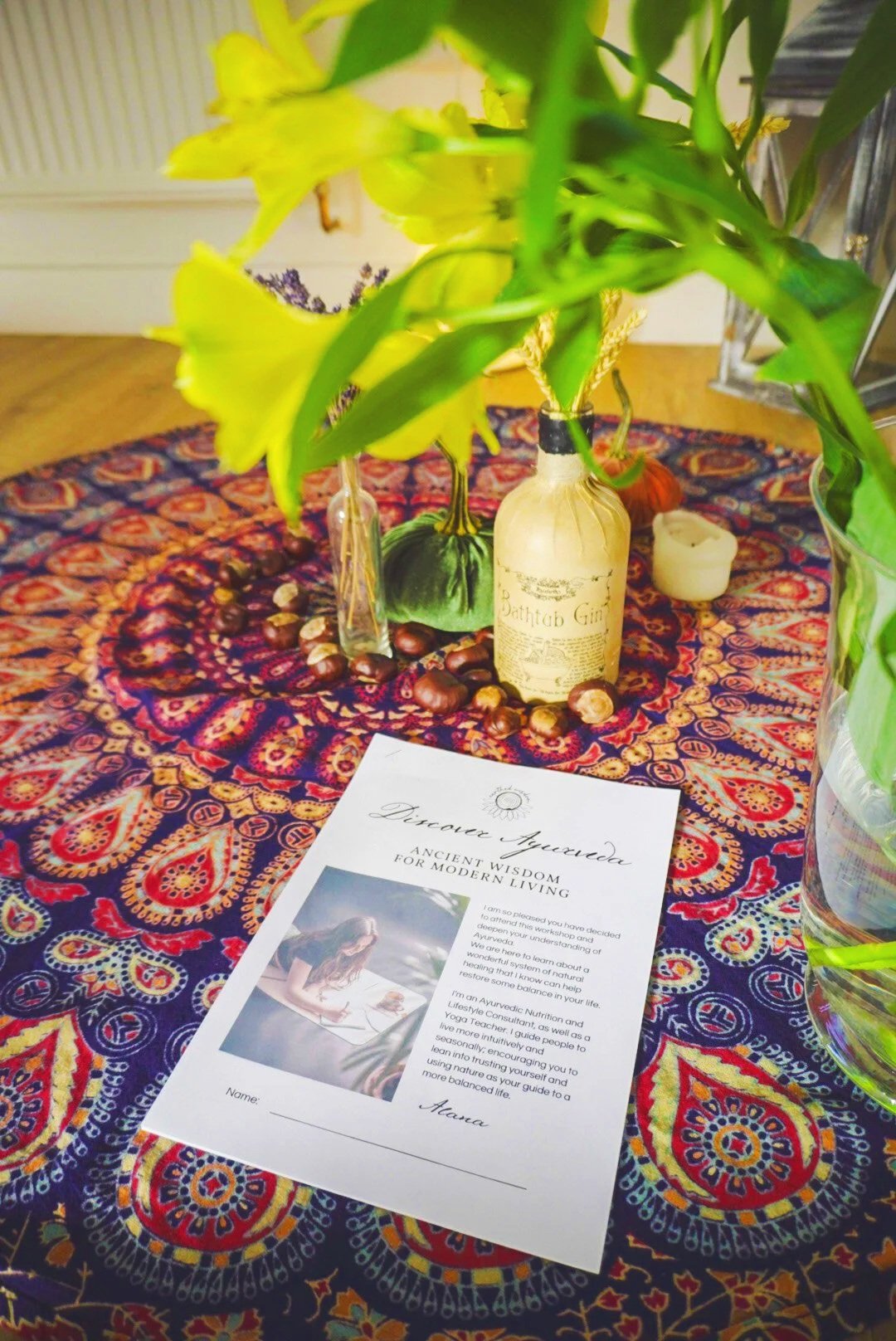 Decorative table cloth on the floor with a colorful paisley design, featuring a white flyer about Ayurvedic wisdom, a glass bowl, small pumpkins, a white candle, and vases with yellow flowers and greenery.