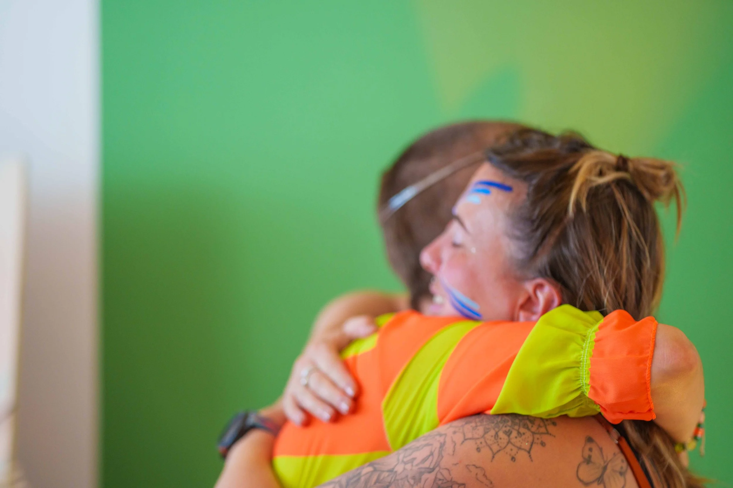 Two people embrace each other in a hug, one with paint on her face and a tattoo on her arm, against a green background.