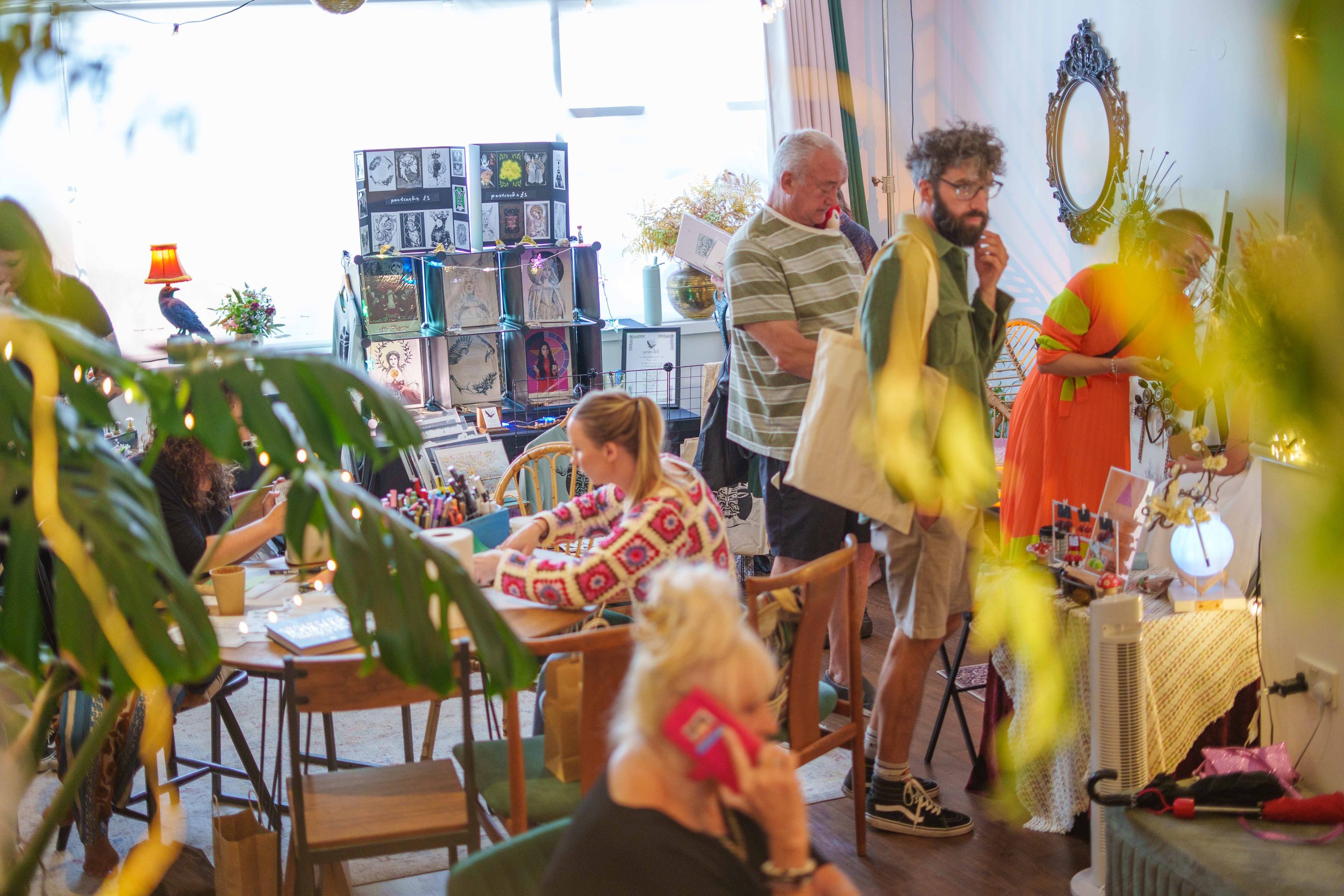 People browsing and shopping at an indoor art and craft market, with tables filled with artwork, crafts, and decorative items, and some displaying framed pictures and artwork.