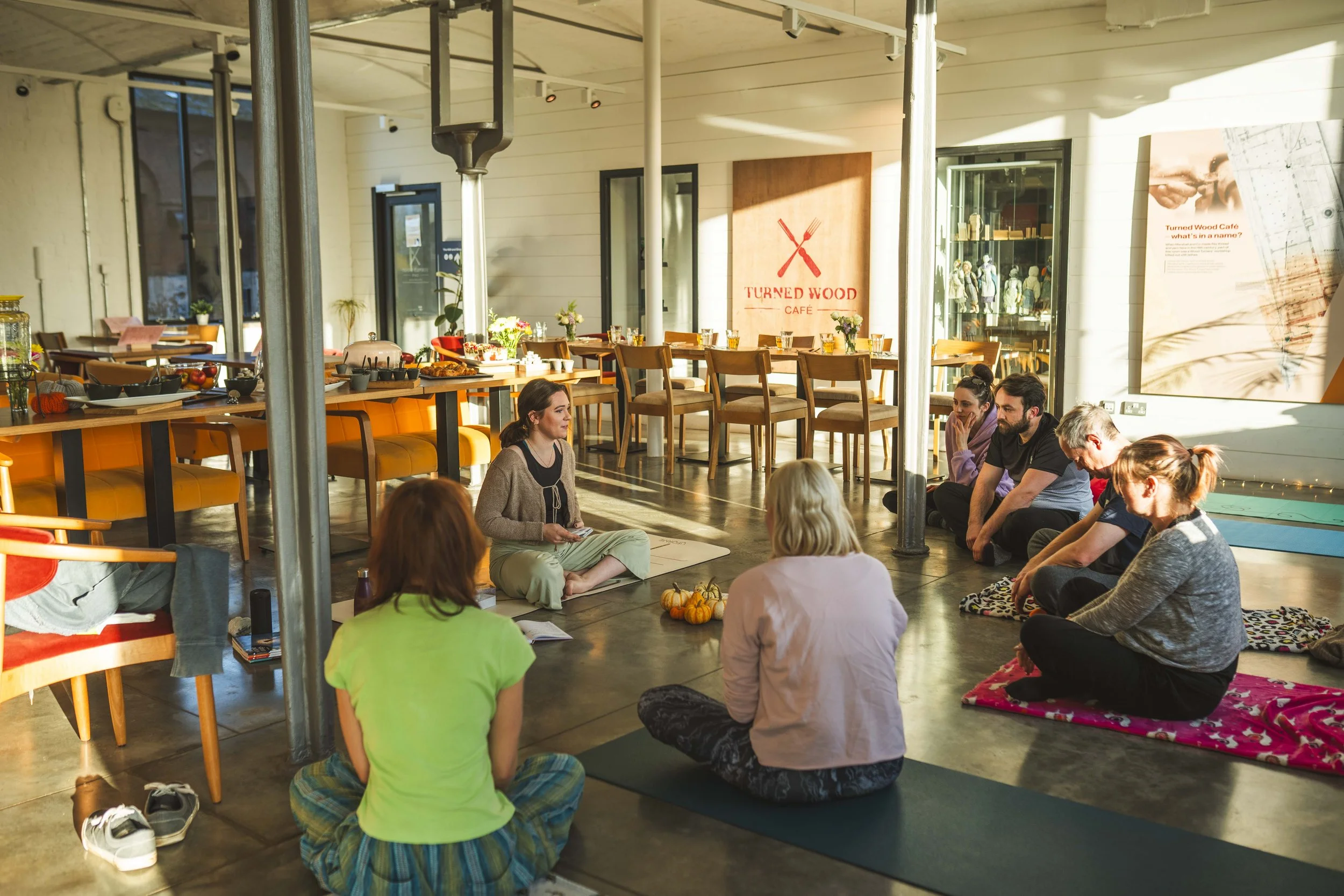 A group of people sitting on yoga mats in a café, participating in a meditation or mindfulness session. The café is decorated with pumpkins and has a sign that reads "Turned Wood Café." The setting is bright with sunlight streaming through large windows.