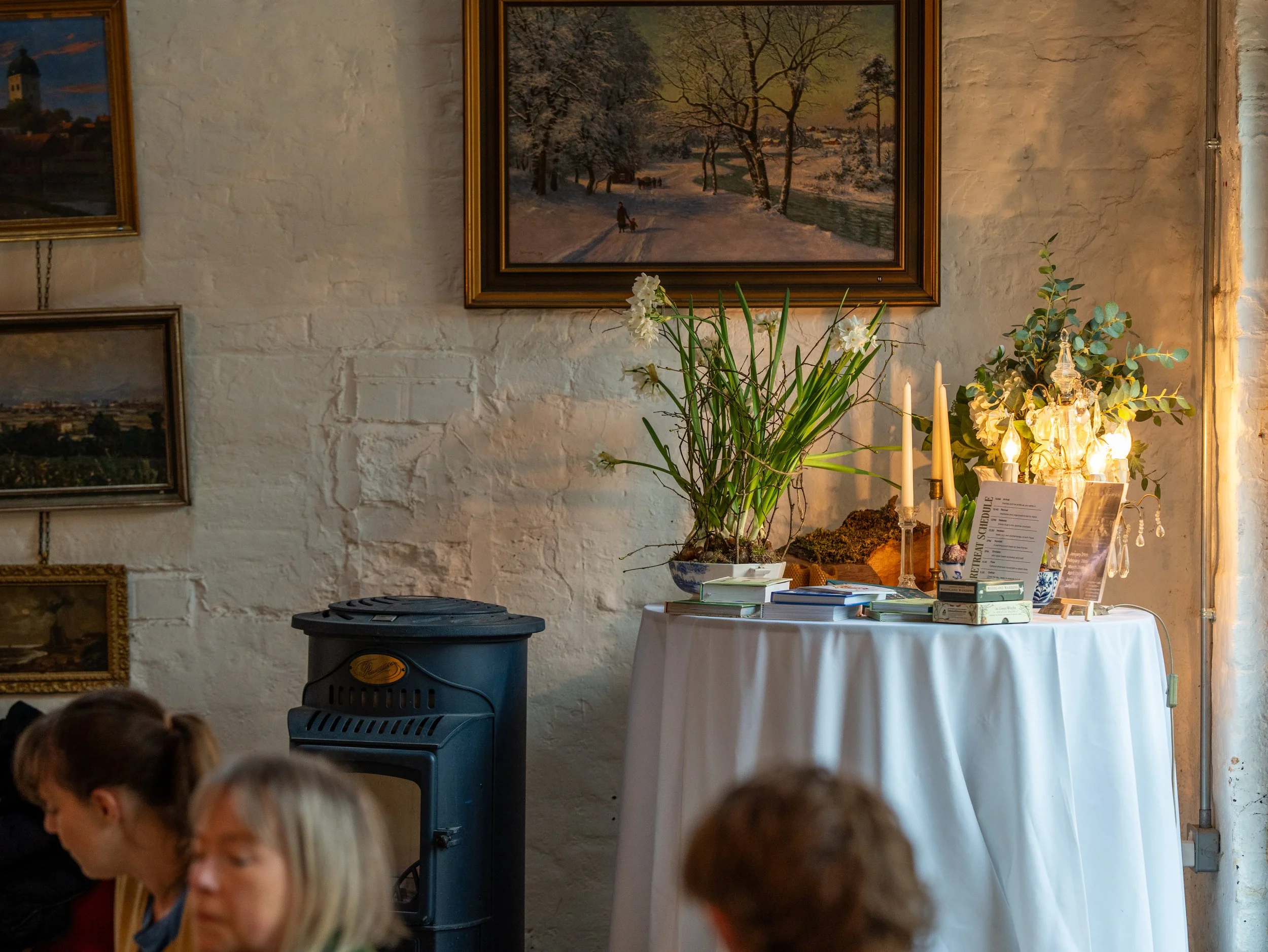 Indoor scene with a white brick wall, framed landscape paintings, a table with white tablecloth, decorative candles, plants, and informational materials, and a small black stove in front of the wall. Partially visible people are in the foreground.