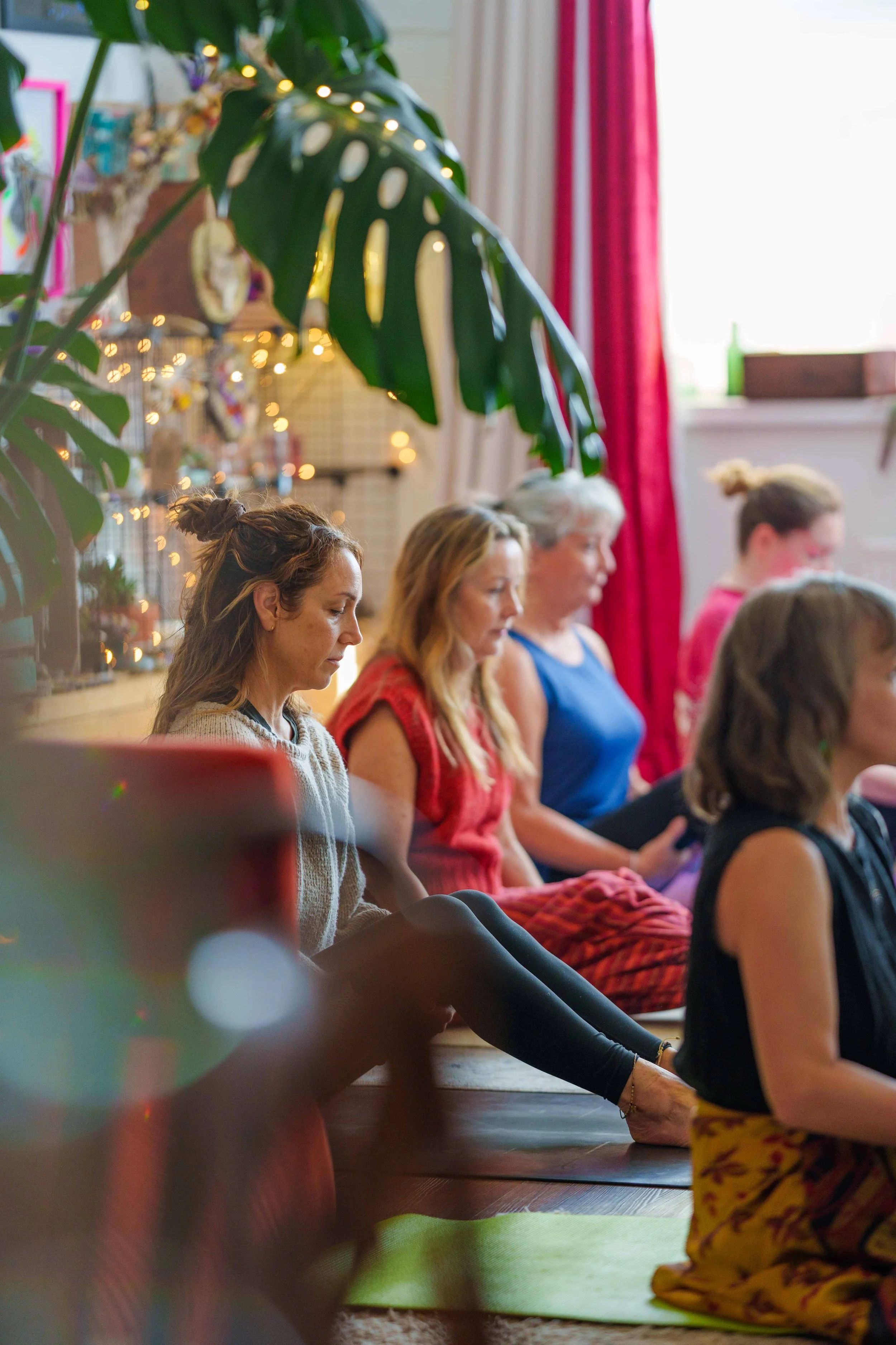 People sitting cross-legged on yoga mats in a meditation practice inside a decorated room with large plant leaves, fairy lights, and red curtains.