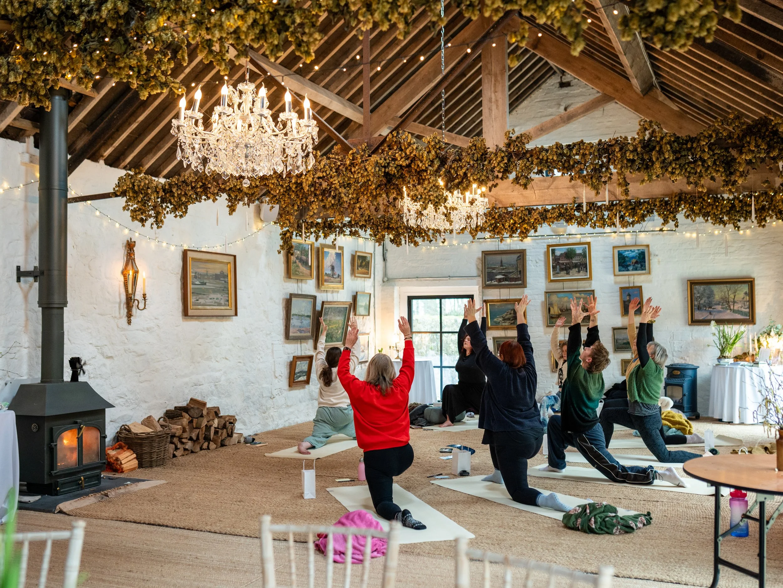 People participating in a yoga class inside a rustic room with white brick walls and framed paintings. The room features a chandelier, dried flowers hanging from the ceiling, and a wood stove. Participants are kneeling on yoga mats with their arms ra