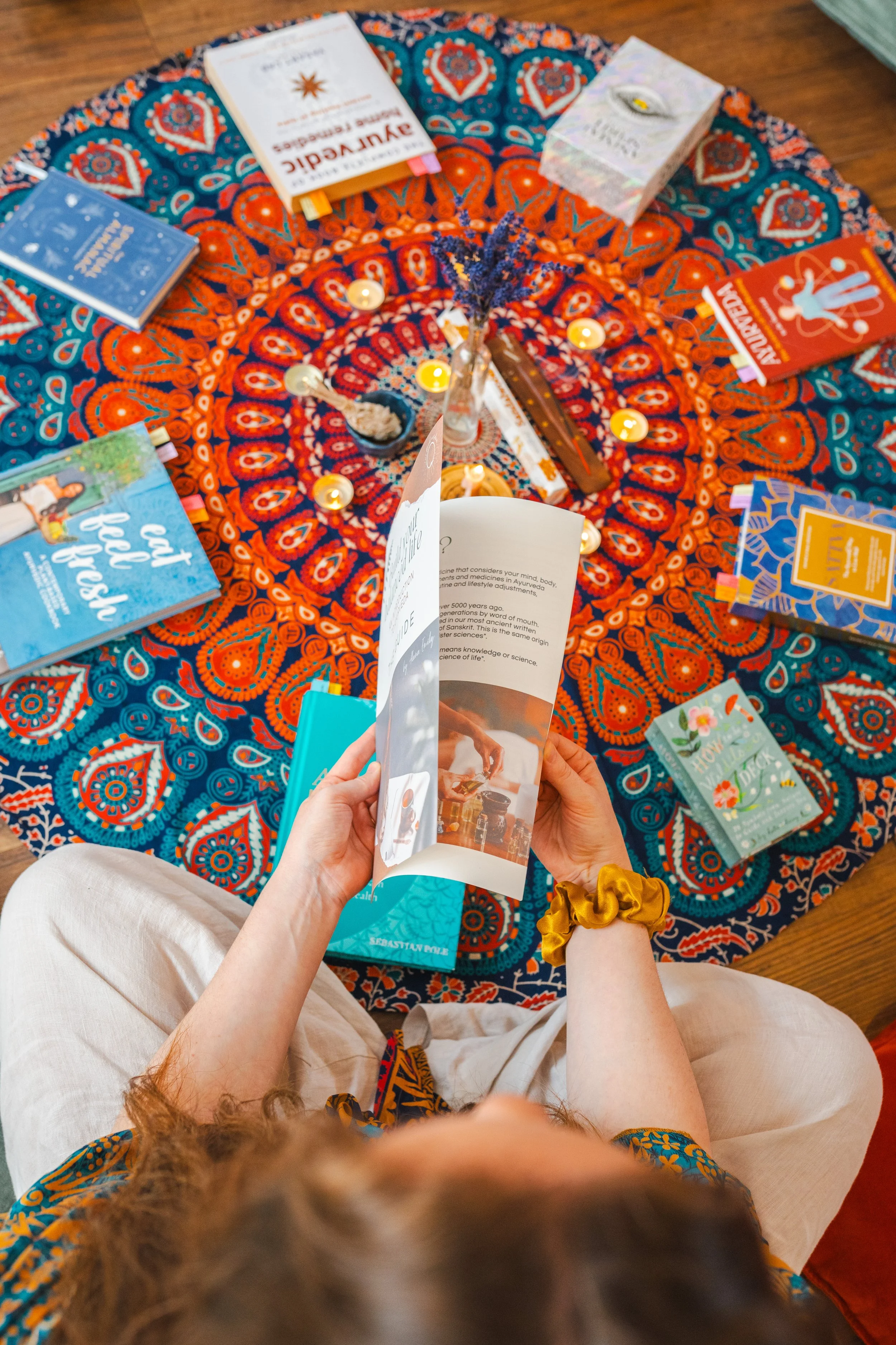 A person sitting on the floor reading a workshop booklet while looking at a circular tablecloth covered with books about Ayurveda. The table has several books, a small vase with dried lavender, some small candles, and other objects on it.