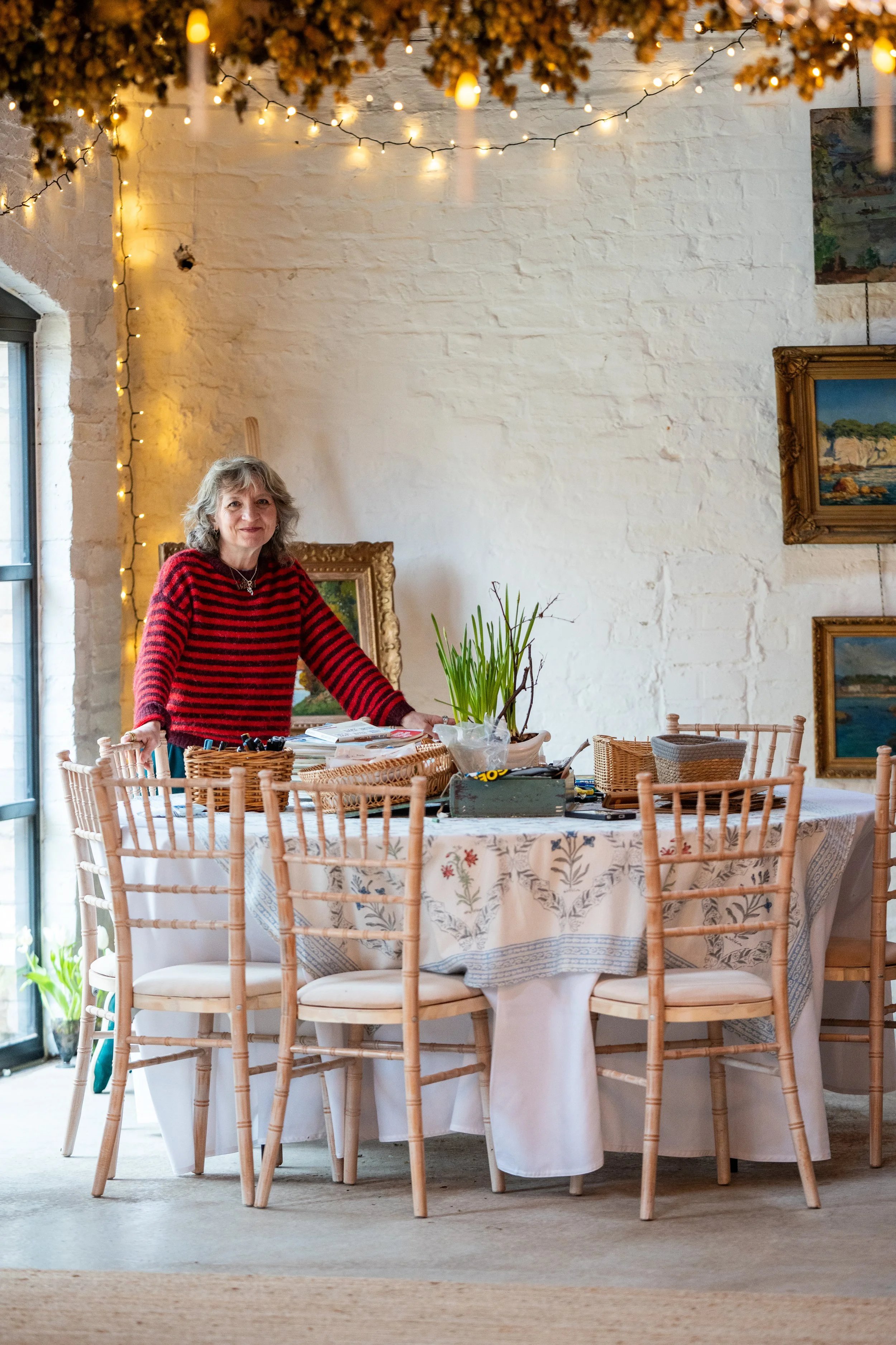 A woman in a red and black striped sweater standing behind a round dining table covered with a floral tablecloth, in a room decorated with string lights and framed paintings on a white brick wall.