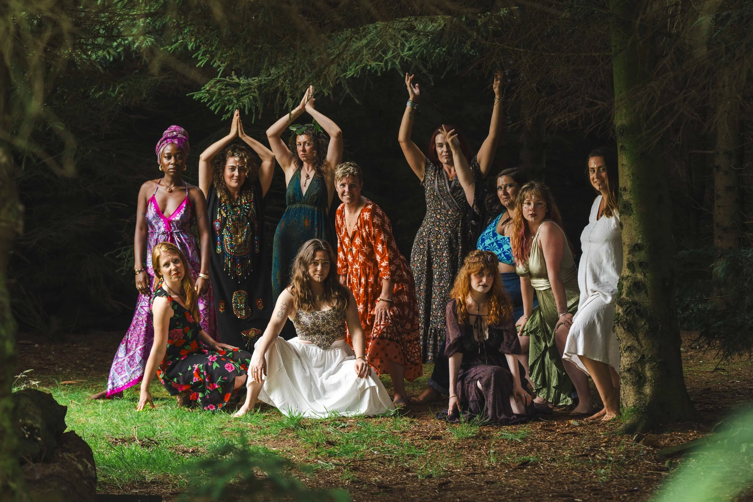 Group of women in colourful dresses posing like goddesses outdoors in a dark wooded area.