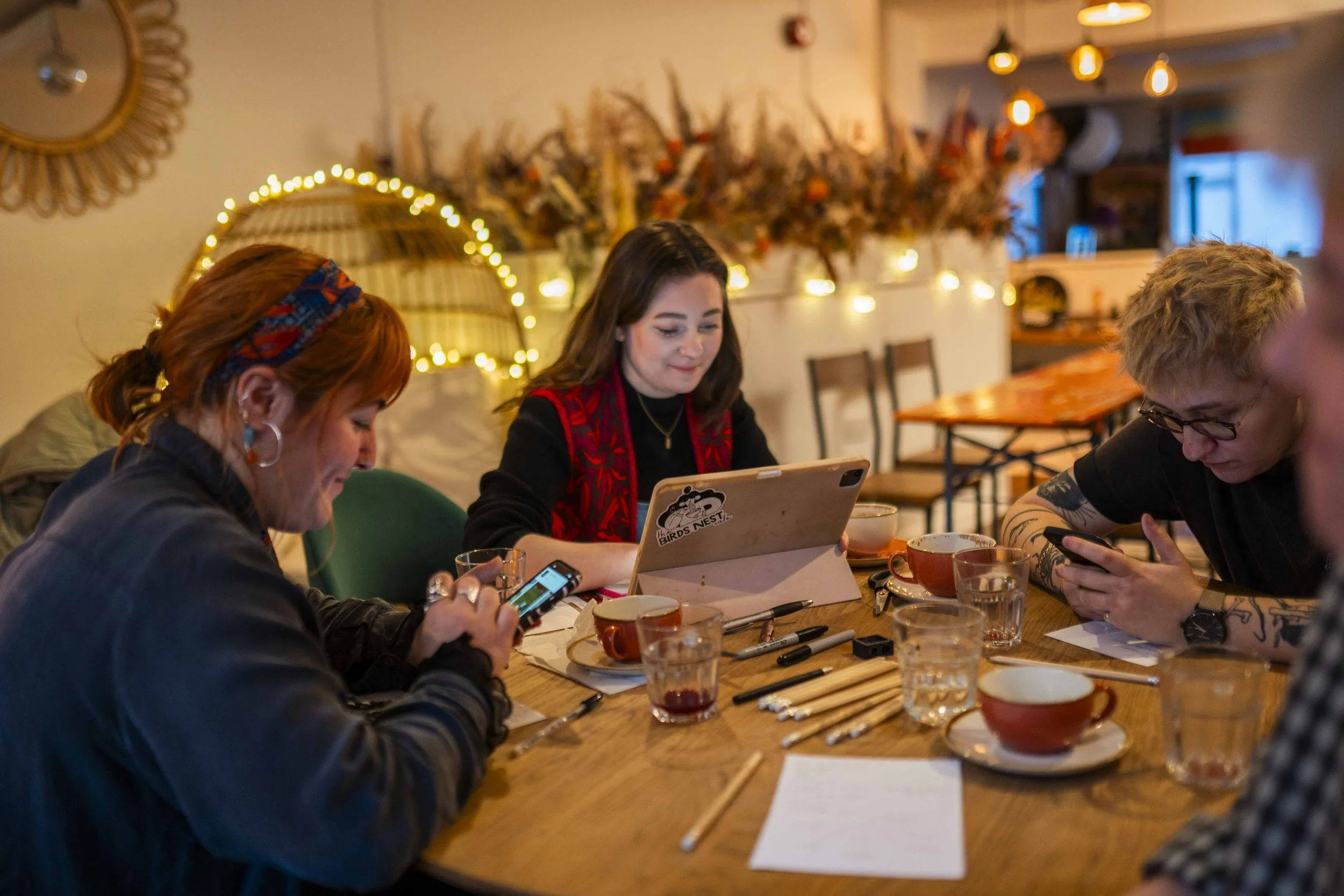 Four people sitting around a wooden table, using phones and a tablet in a cozy room decorated with warm lights and dried flowers in the background.