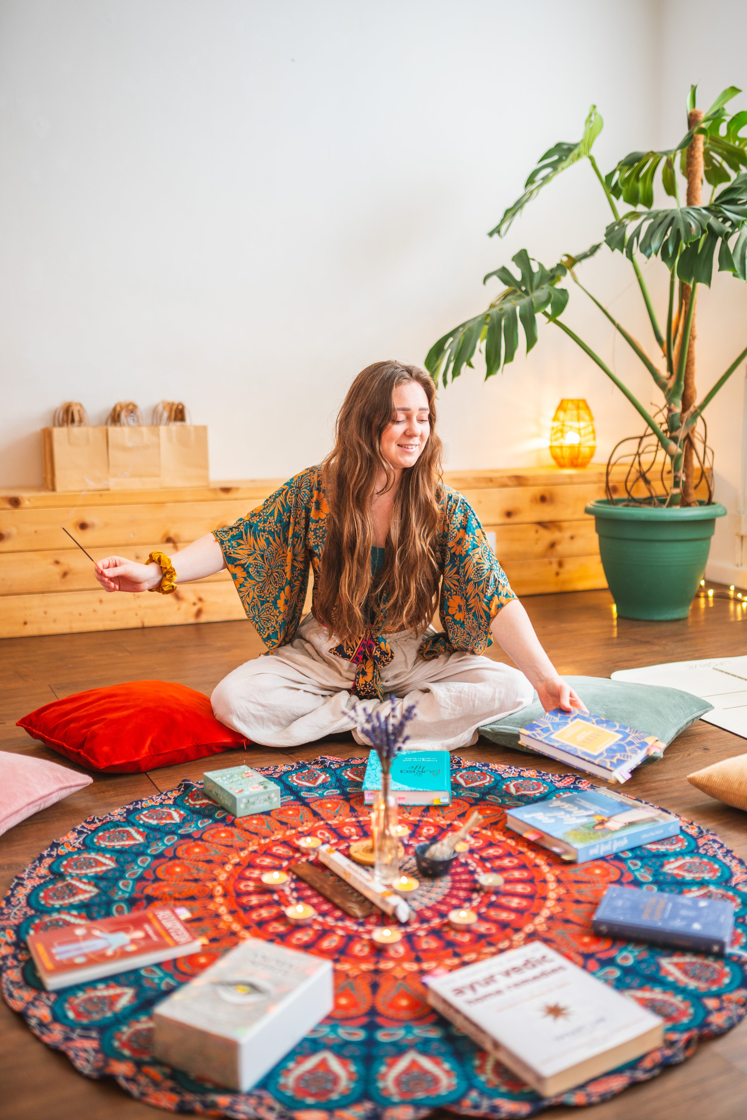 A woman with long brown hair sitting on a red pillow on a wooden floor, surrounded by books and candles, in a cosy room with a large green potted plant, warm lighting, and a decorated round cloth on the floor.