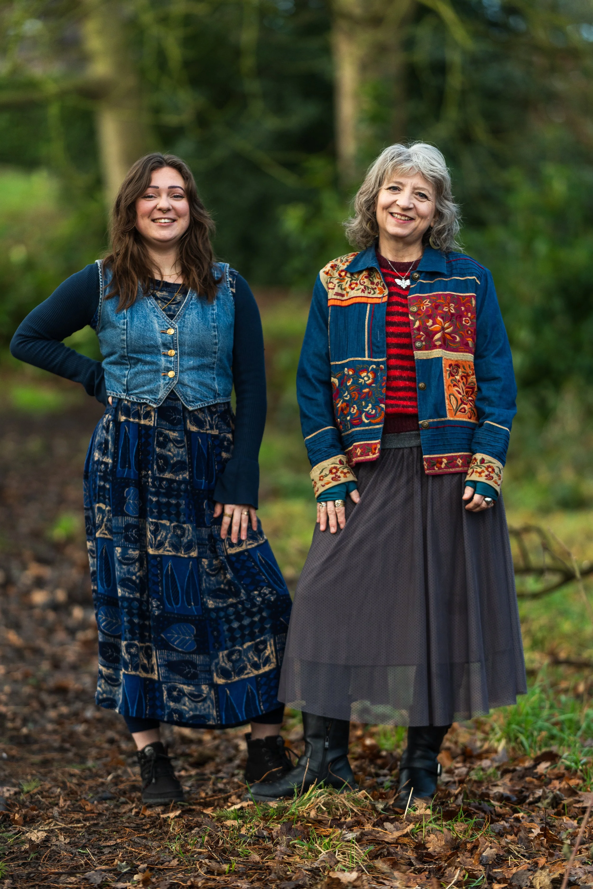 Two women standing in a wooded outdoor setting, smiling at the camera. One appears to be middle-aged with gray hair, wearing a colorful embroidered denim jacket, striped red shirt, and a long dark skirt. The other is younger with long brown hair, wearing a denim vest over a long patterned dress.
