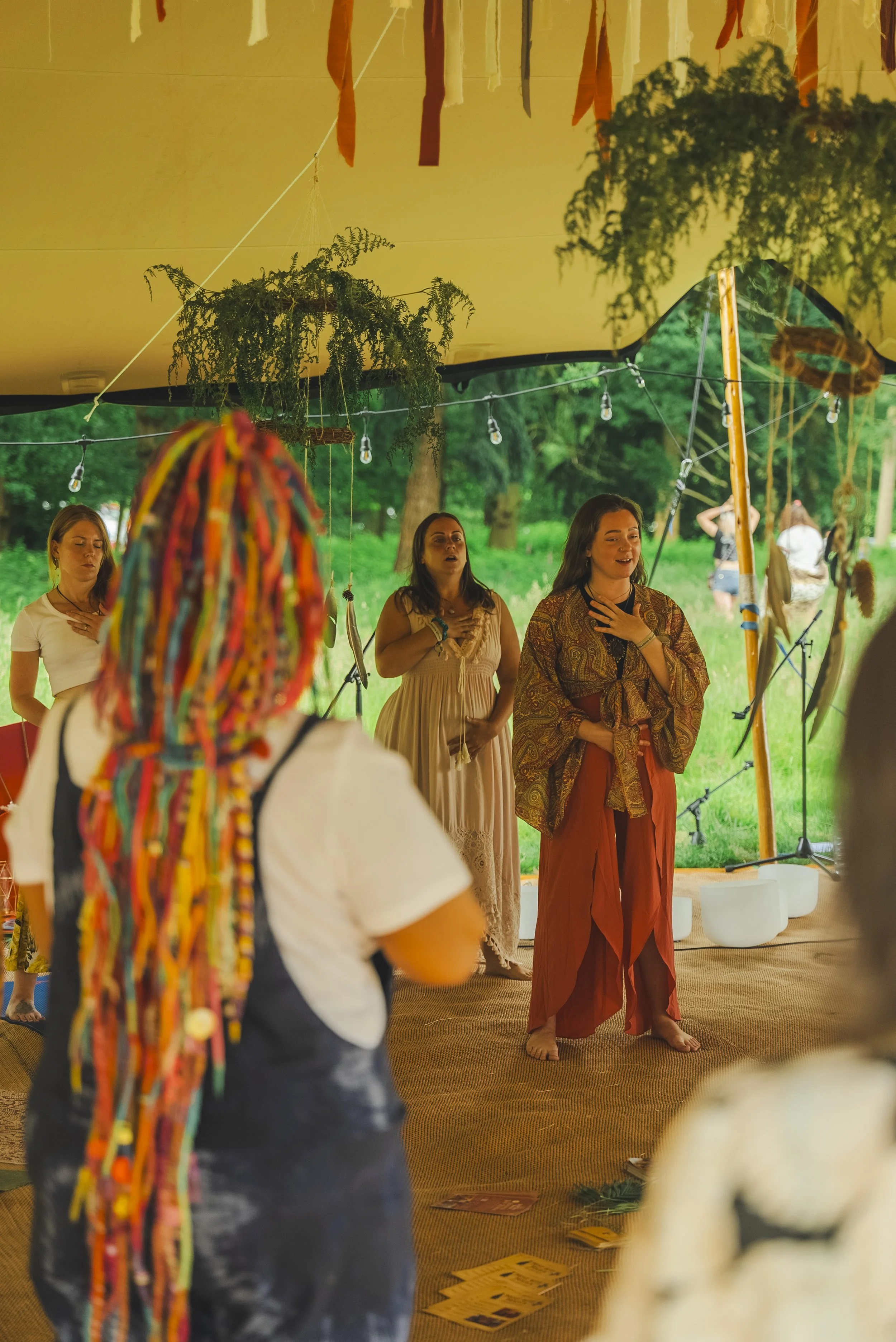 Group of women inside a decorated tent, some standing with hands on chest, participating in a spiritual or meditative gathering, with green outdoor scenery visible through open sides.