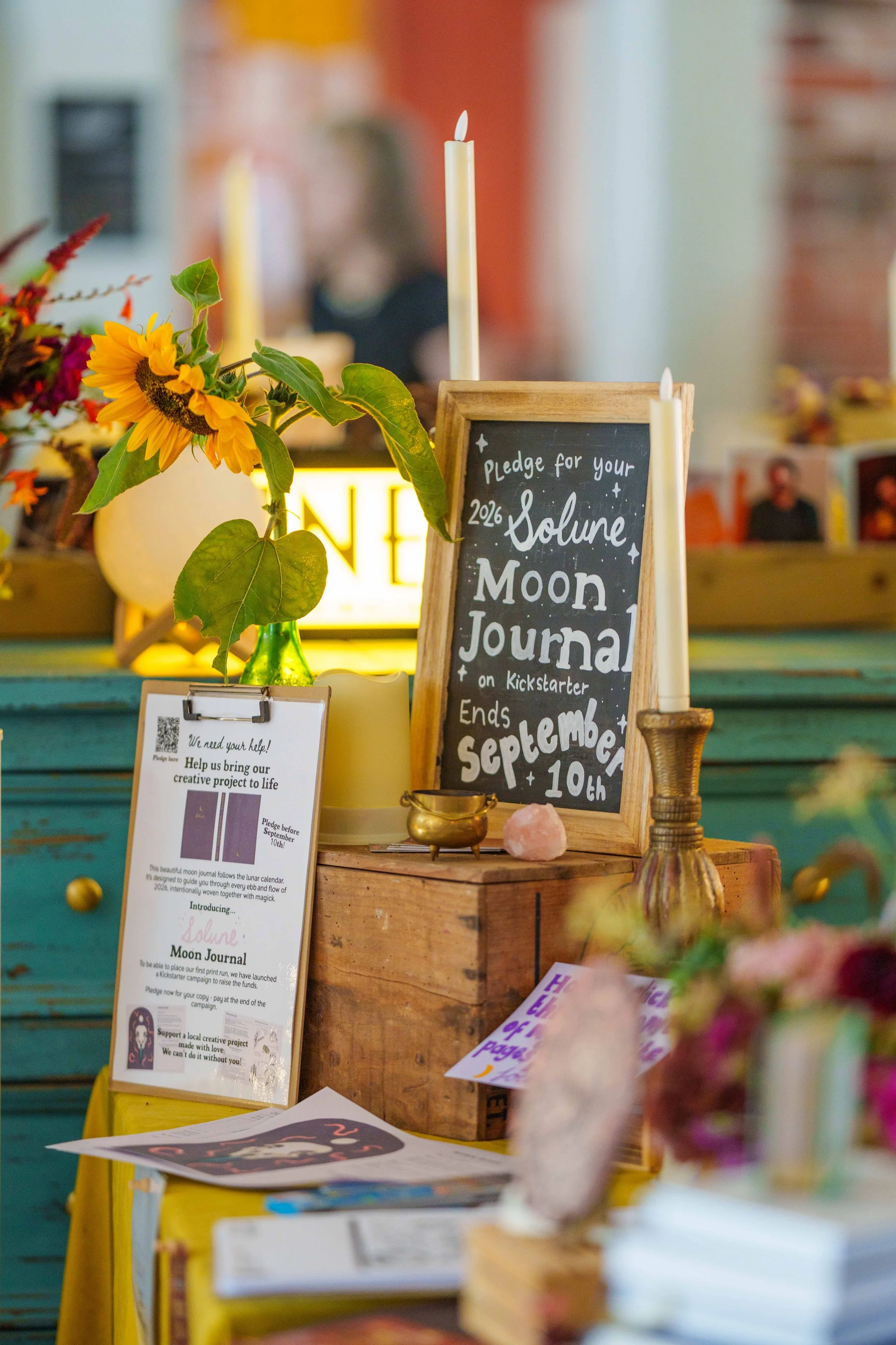 Sign promoting the Solune Moon Journal Kickstarter, with candles, flowers, and informational flyers on a yellow table.