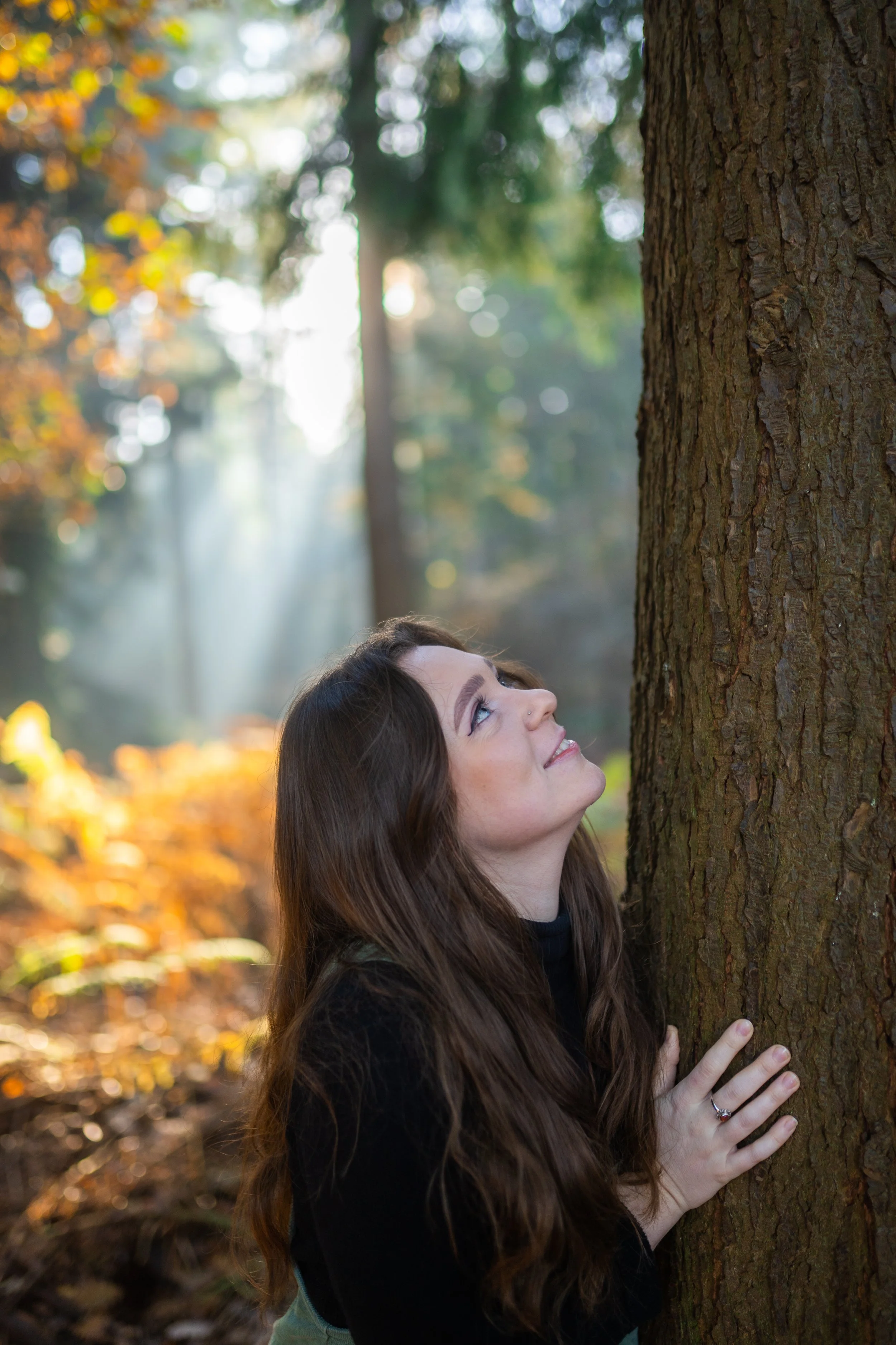 A woman with long, wavy brown hair smiling and looking up while hugging a tree in a forest with sunlight filtering through autumn-colored leaves.