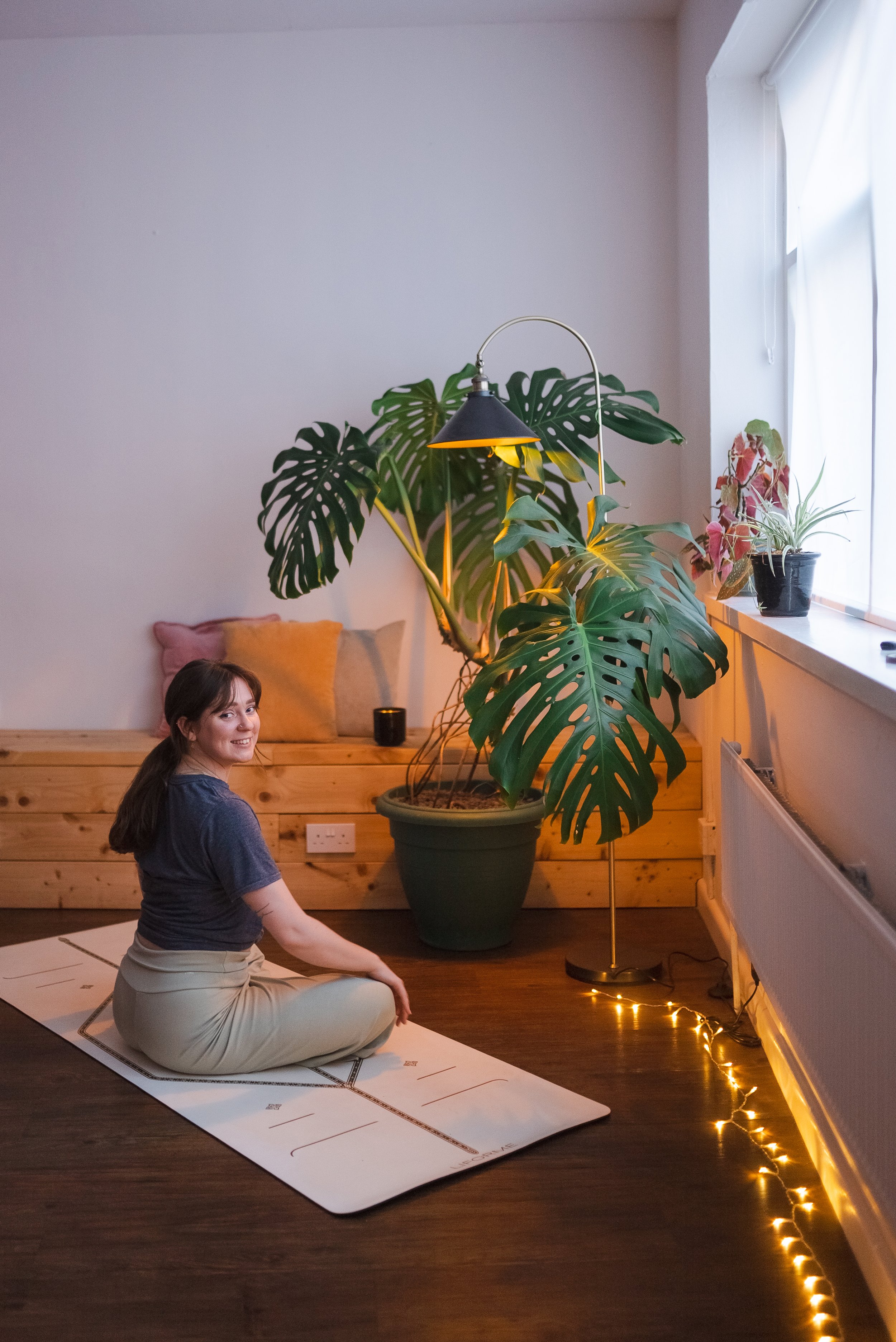 A woman sitting on a yoga mat in a yoga pose indoors near a large potted monstera plant, with string lights on the floor, potted plants on the windowsill, and a colorful cushion on a wooden bench.